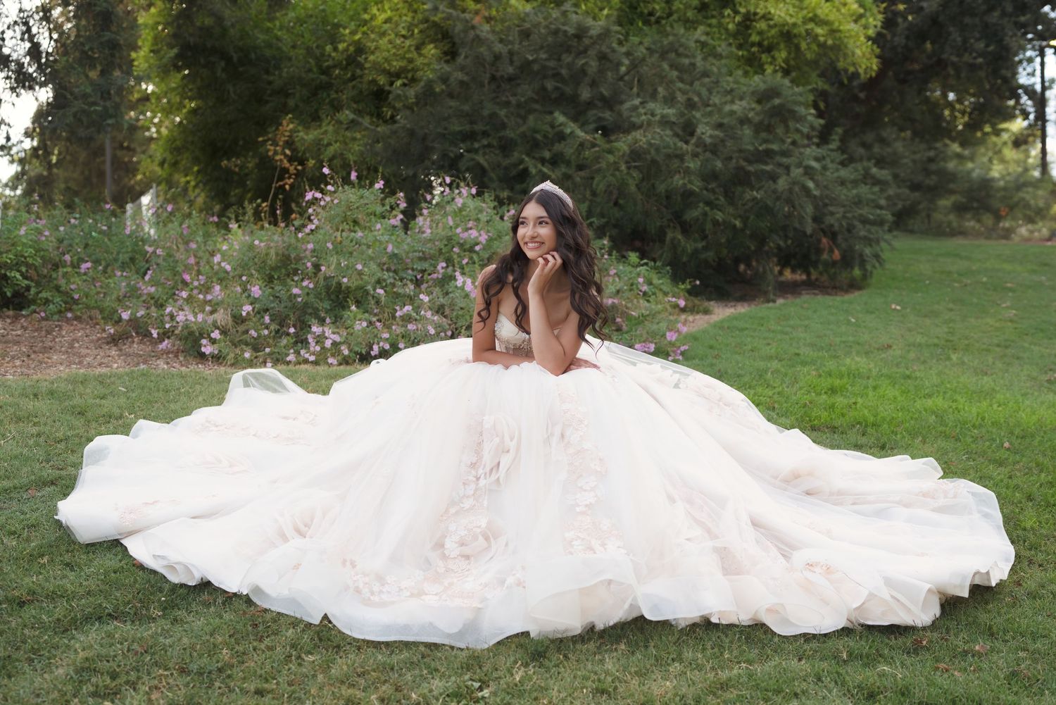 Bride in a voluminous blush ball gown with floral details, sitting on green grass in a garden setting.