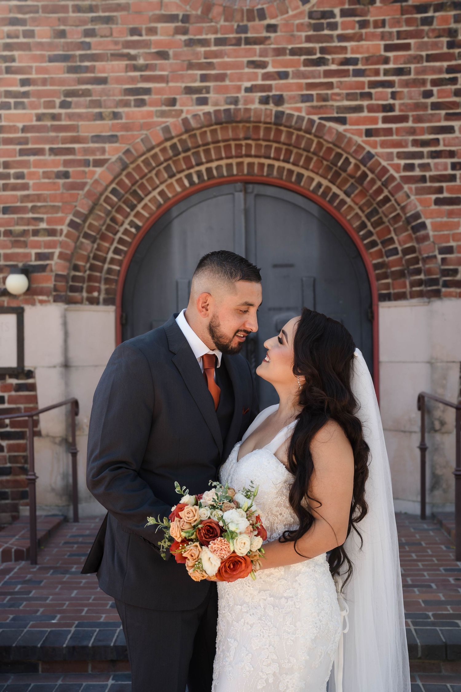 Bride and groom share a romantic moment in front of a brick arch doorway, bride holding a bouquet of orange and white flowers.