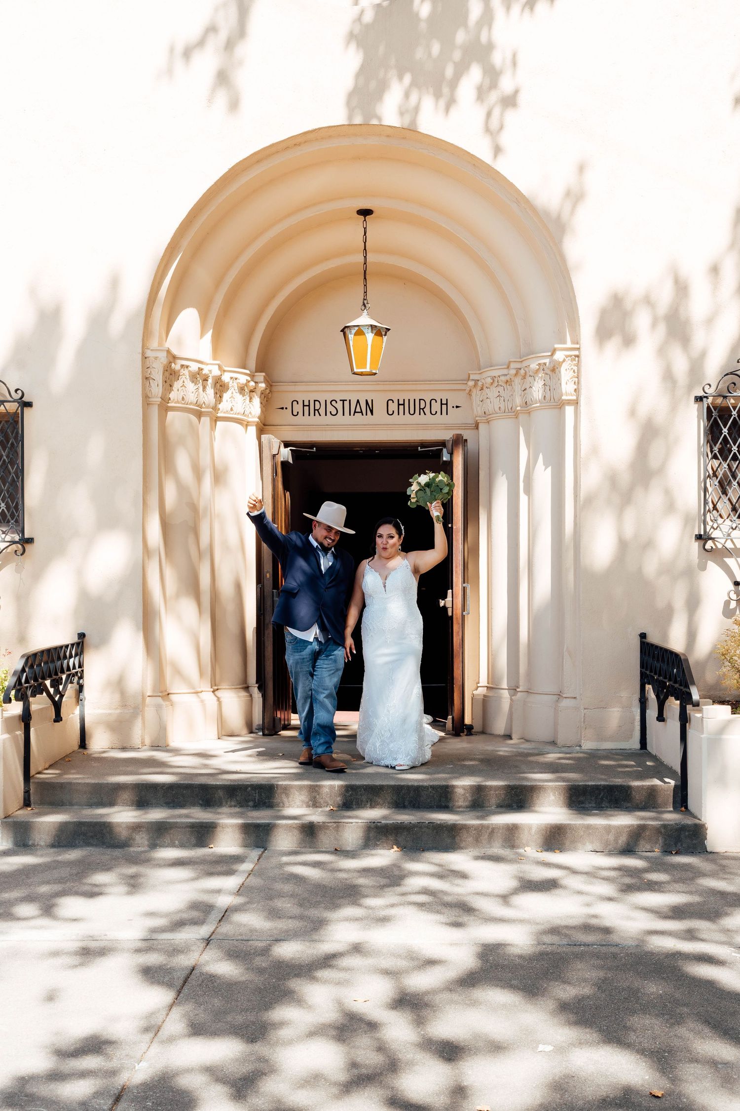 Joyful newlyweds raise arms outside Christian Church entrance after ceremony.