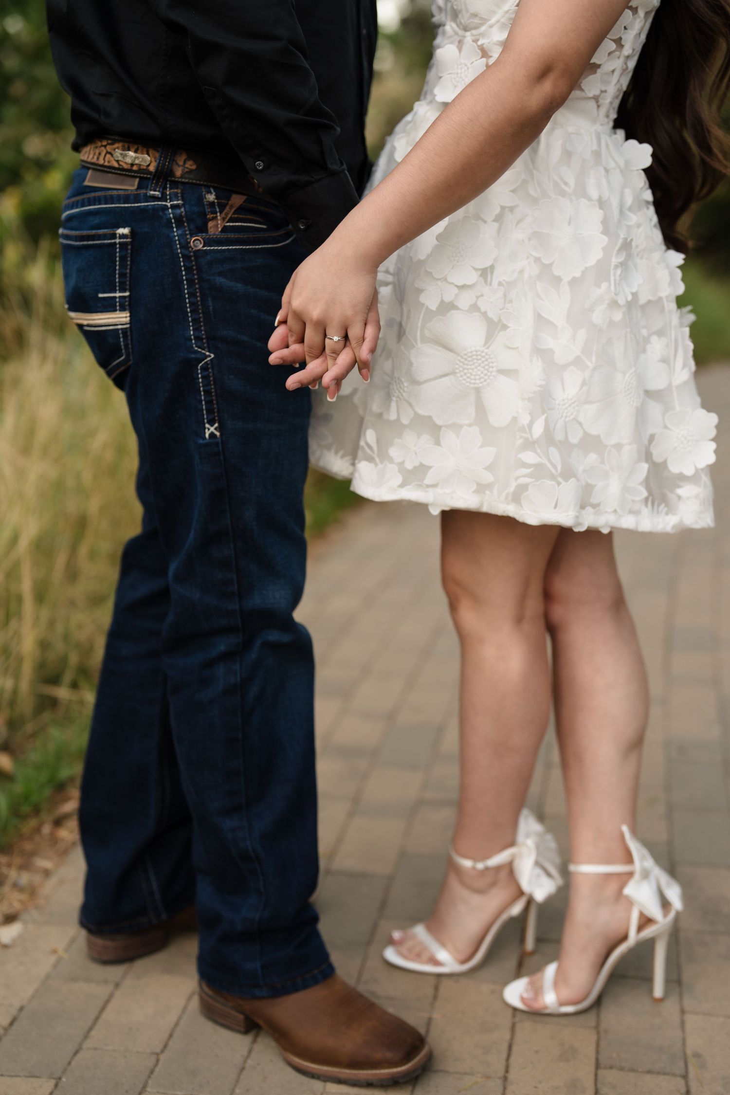 Close-up of couple holding hands showing engagement ring, man in cowboy boots and jeans, woman in white heels.