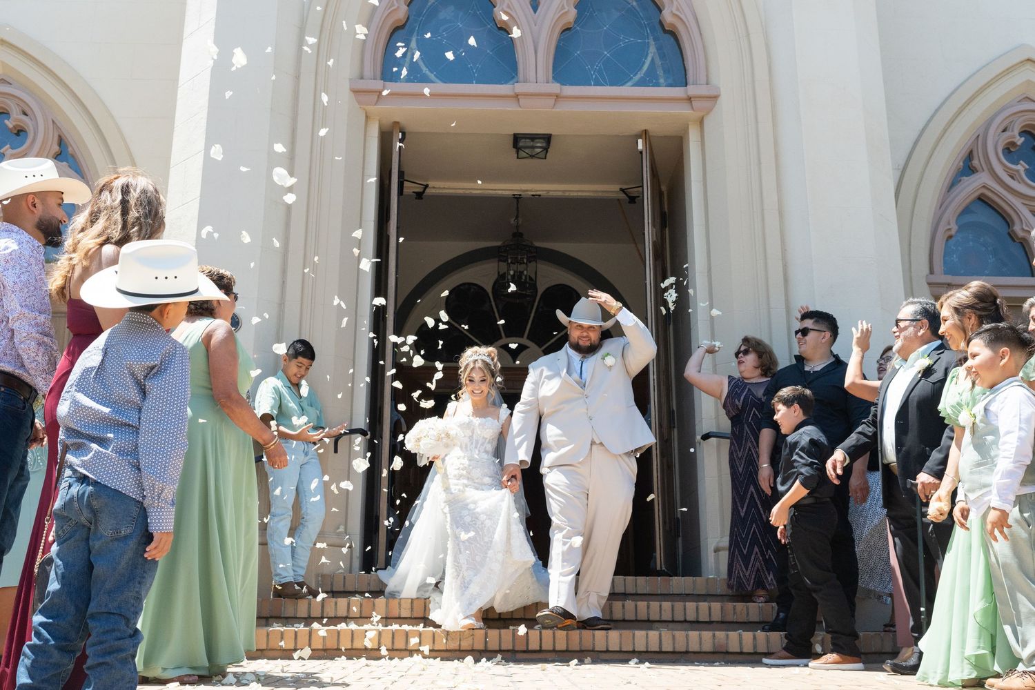 Bride and groom exiting a church as guests toss confetti, groom in white suit and cowboy hat.