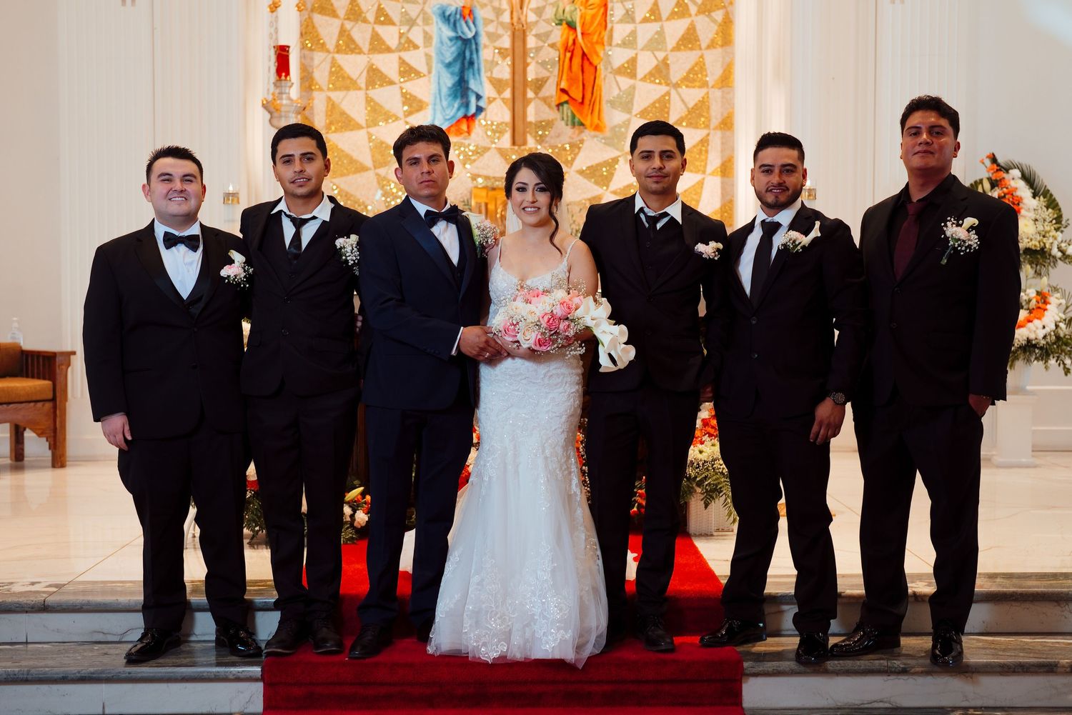 Bride in white gown and groom in black tuxedo pose with groomsmen inside a decorated church.