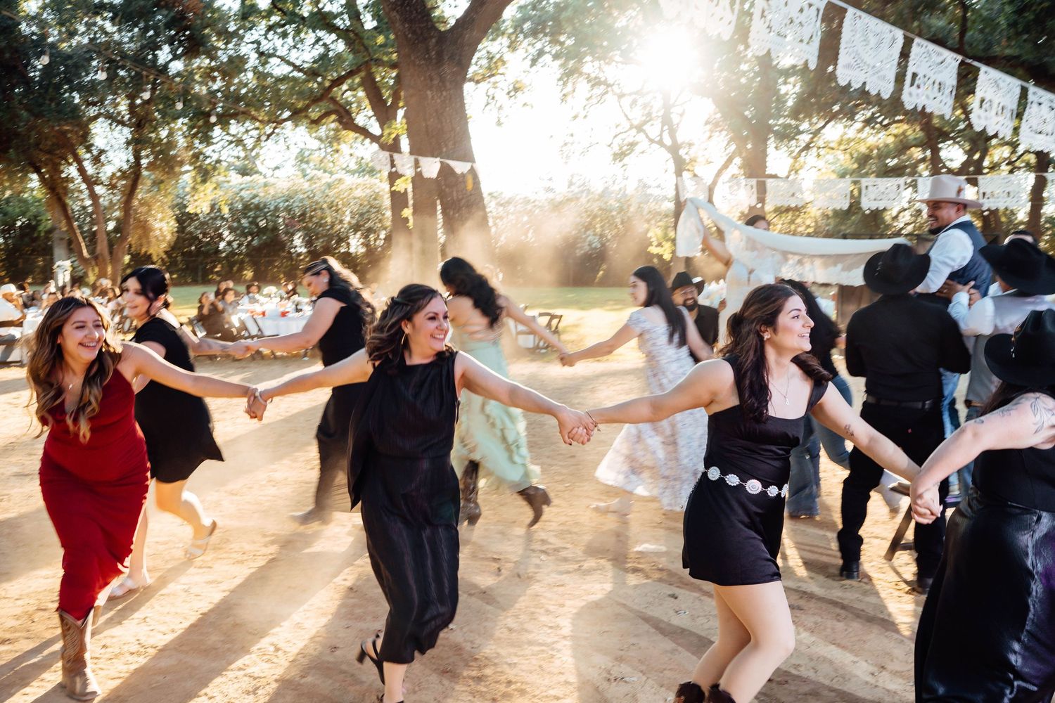Wedding guests hold hands and dance in a chain line at a sunny outdoor rustic reception.