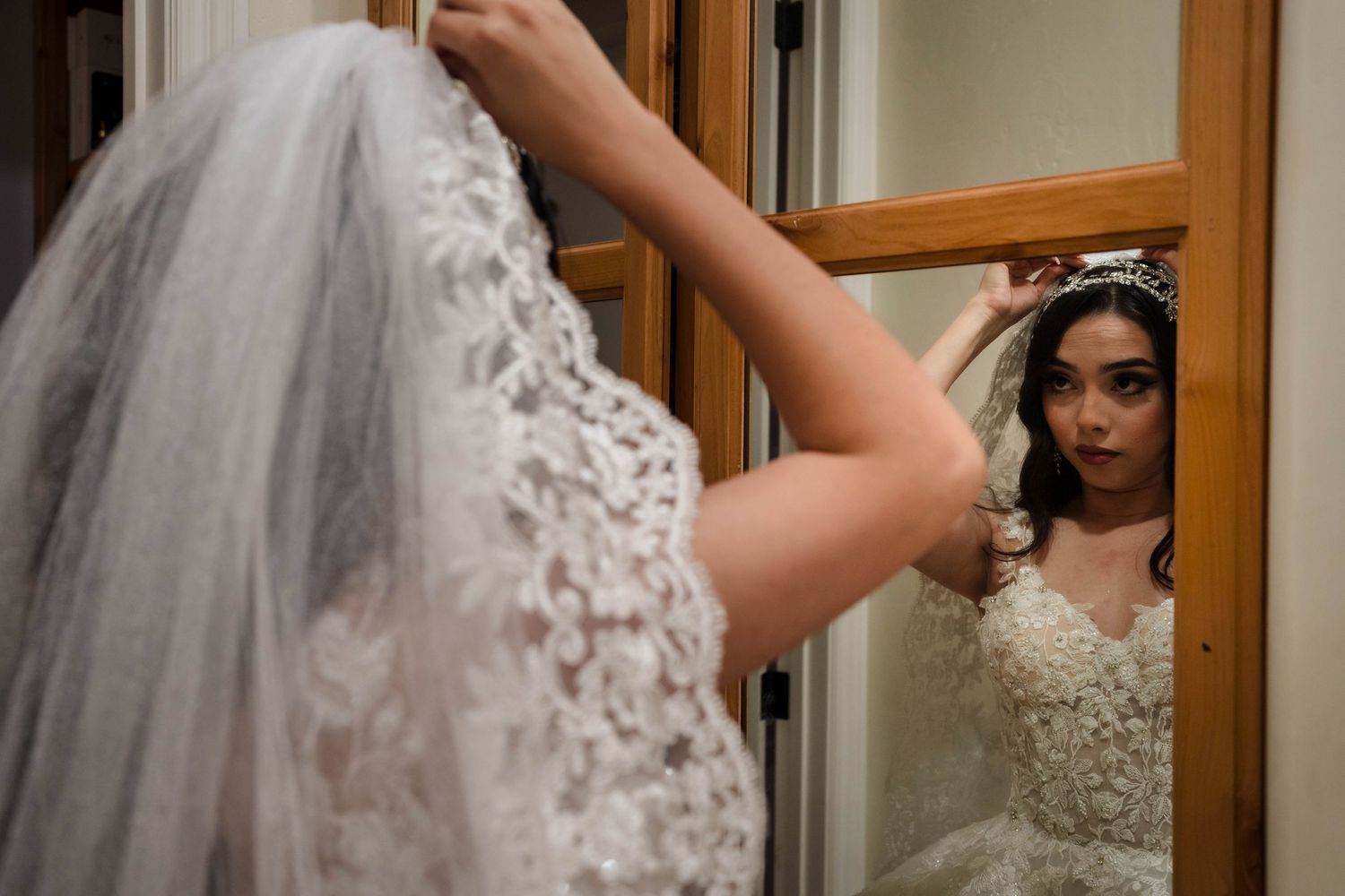 Bride in lace wedding gown and tiara adjusting veil while looking in mirror during bridal preparation.