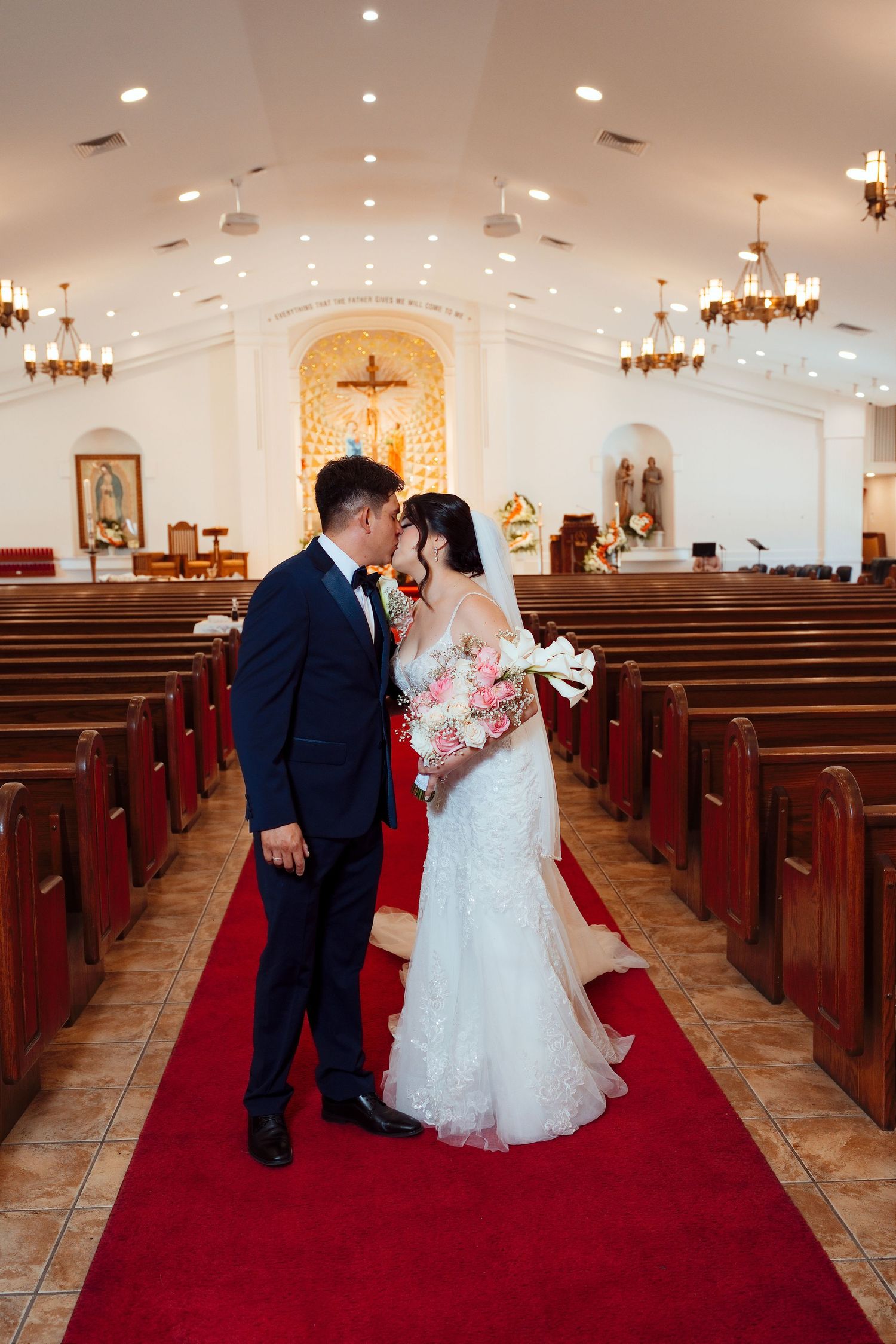 Bride and groom share a kiss on red aisle of white church, bride holding pink and white bouquet in lace gown.
