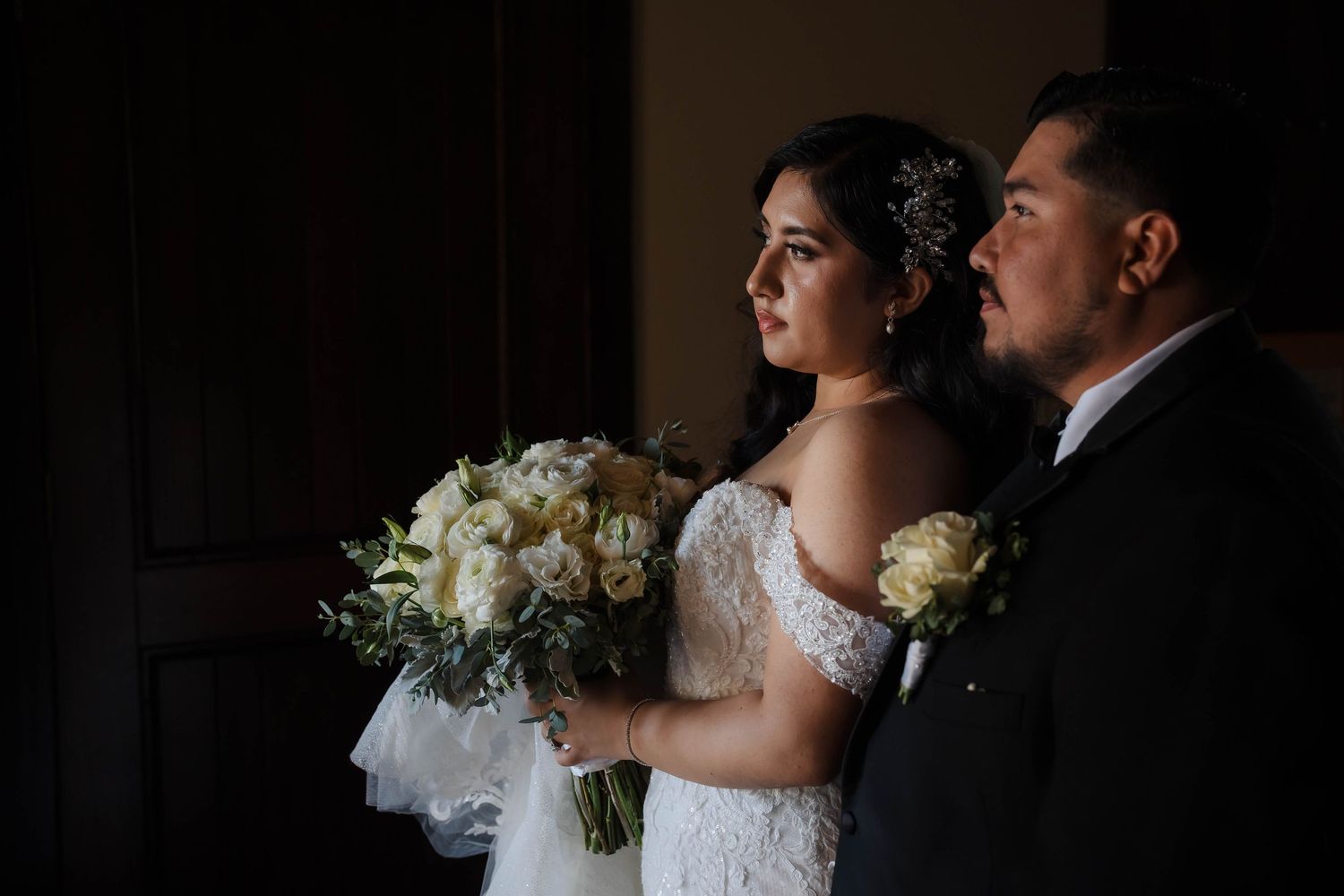 Bride in white lace gown holding bouquet of white roses stands beside groom in black suit in elegant wedding portrait.