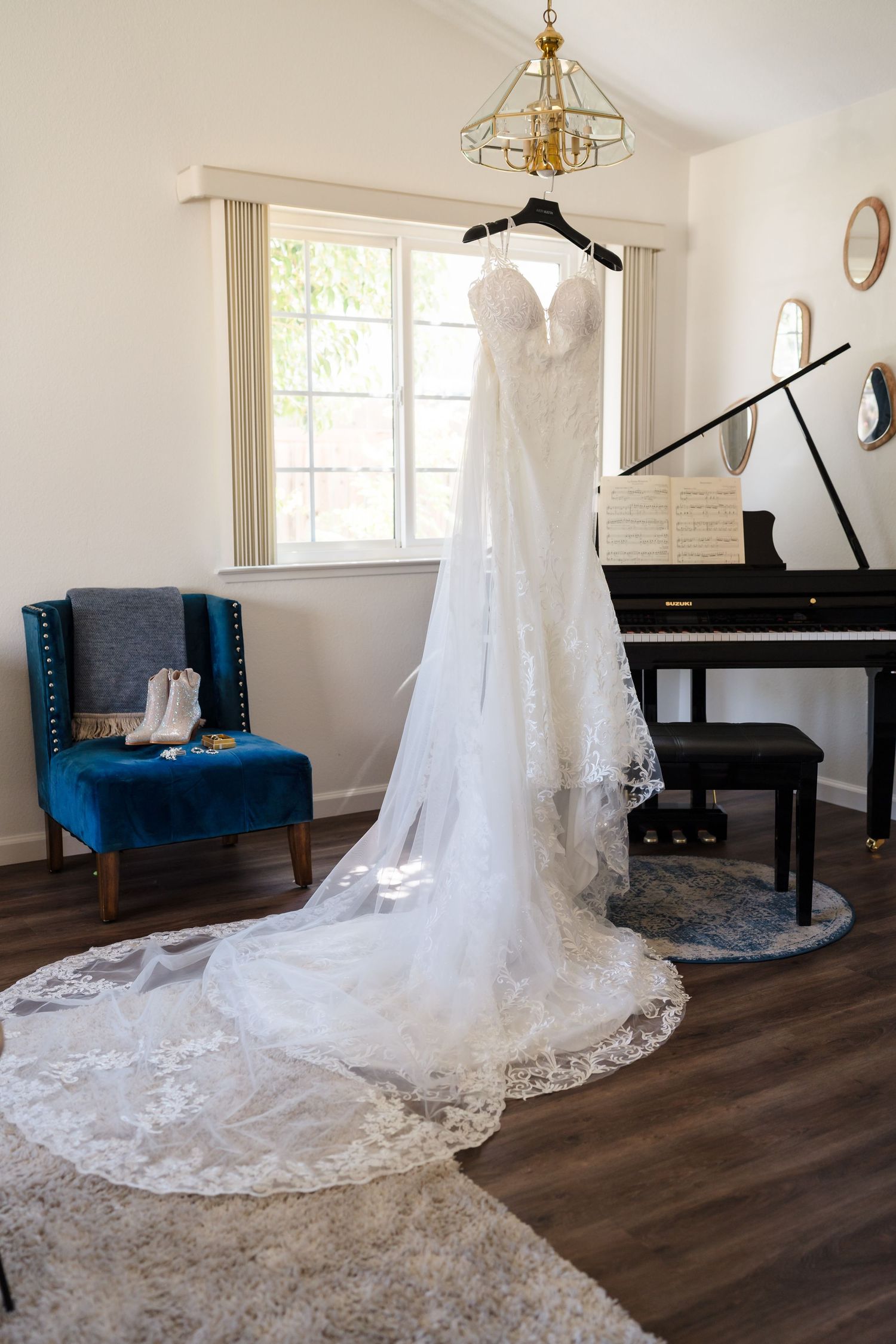White lace wedding dress with long train hanging in elegant room with grand piano and blue velvet chair.