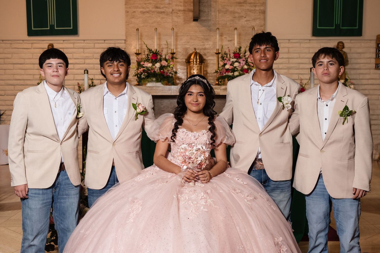 A quinceañera girl in a pink ball gown poses with four young men in tan blazers inside a decorated church.