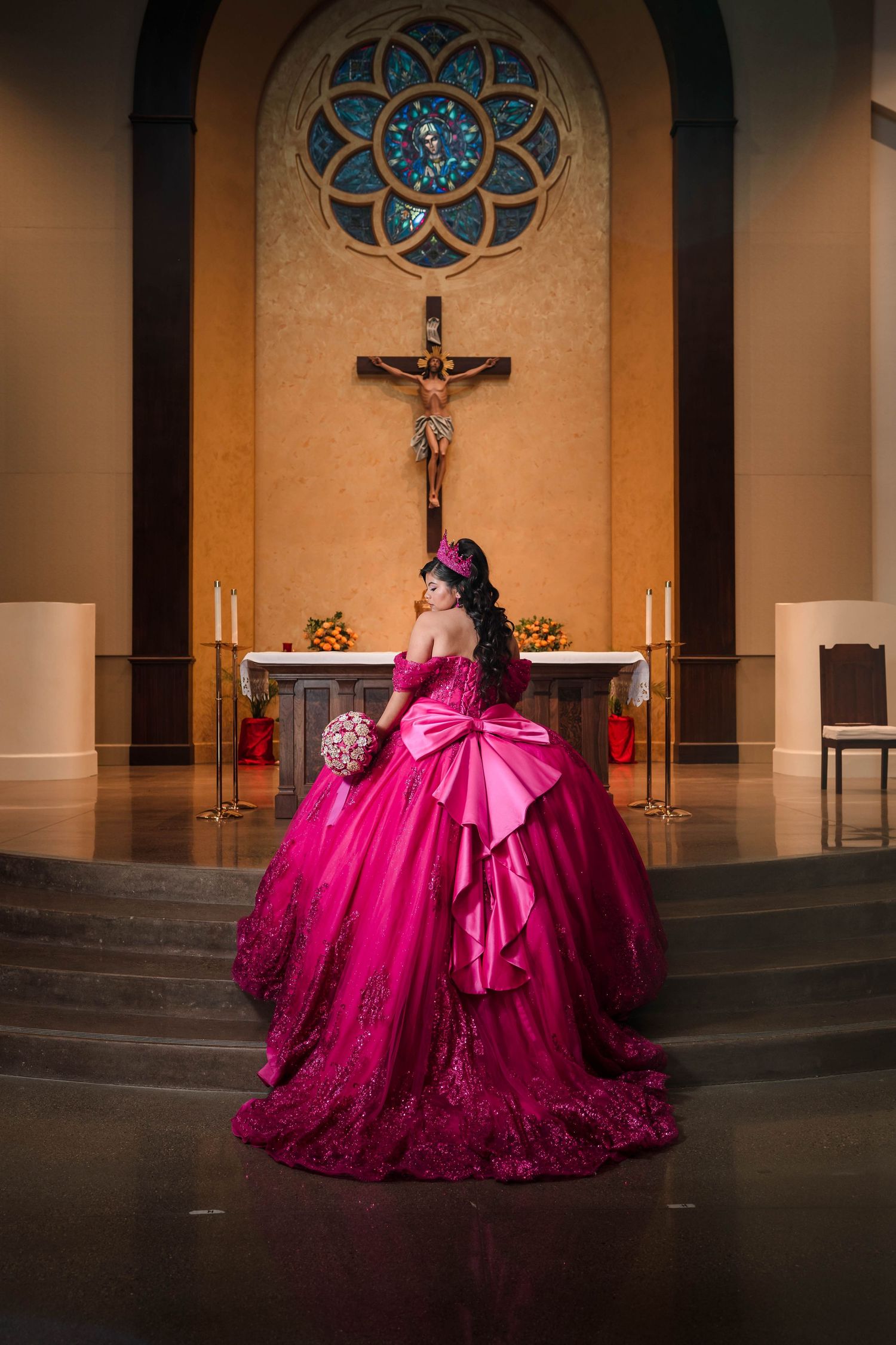 Quinceañera girl in elaborate hot pink ball gown kneels before church altar with crucifix backdrop.