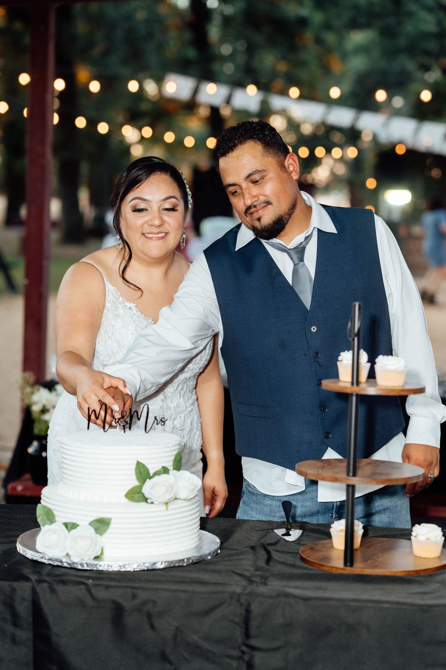 Bride and groom cutting their two-tier white wedding cake decorated with roses at reception.