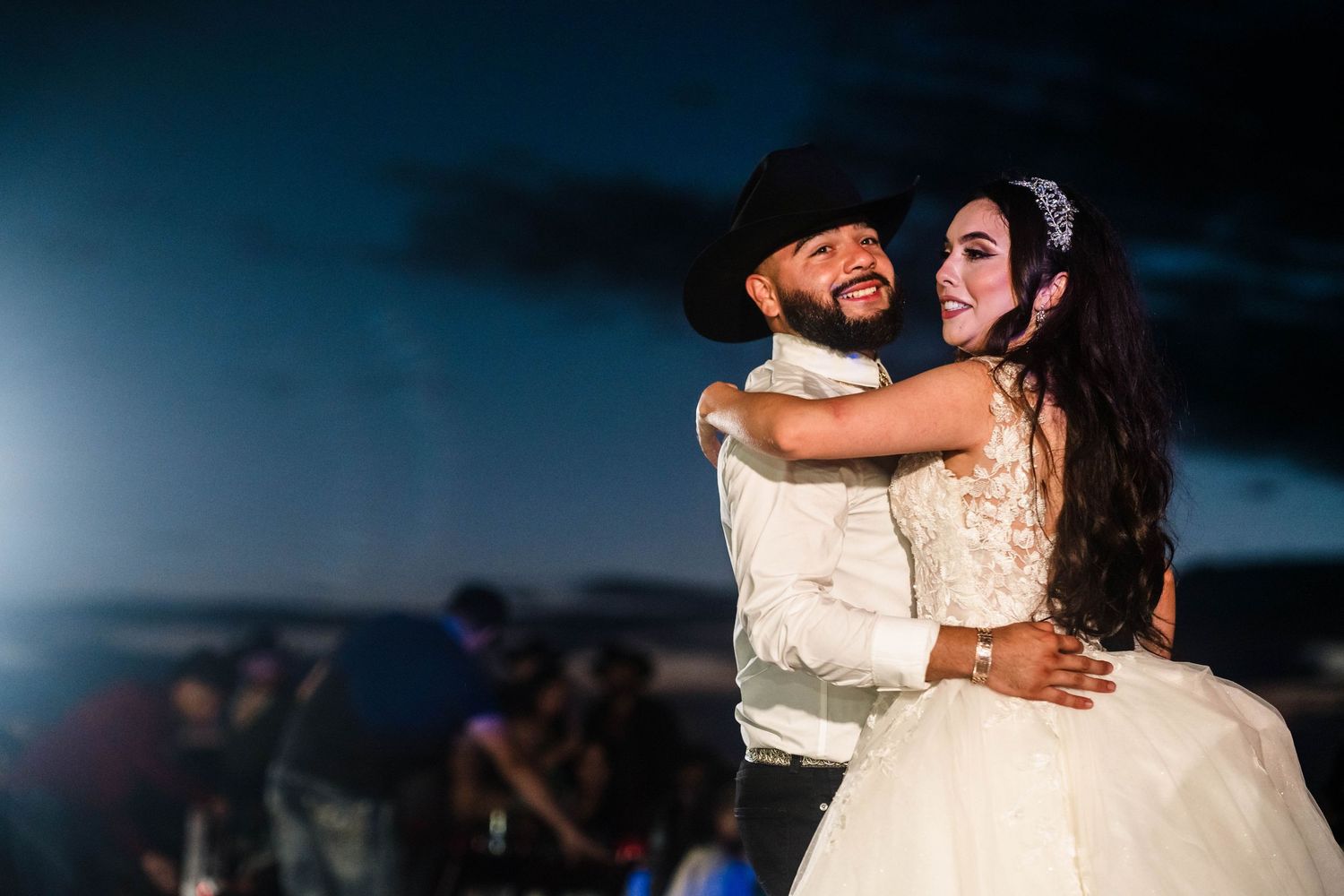 Couple sharing first dance at outdoor wedding reception under night sky, groom in black cowboy hat embracing bride in white gown.