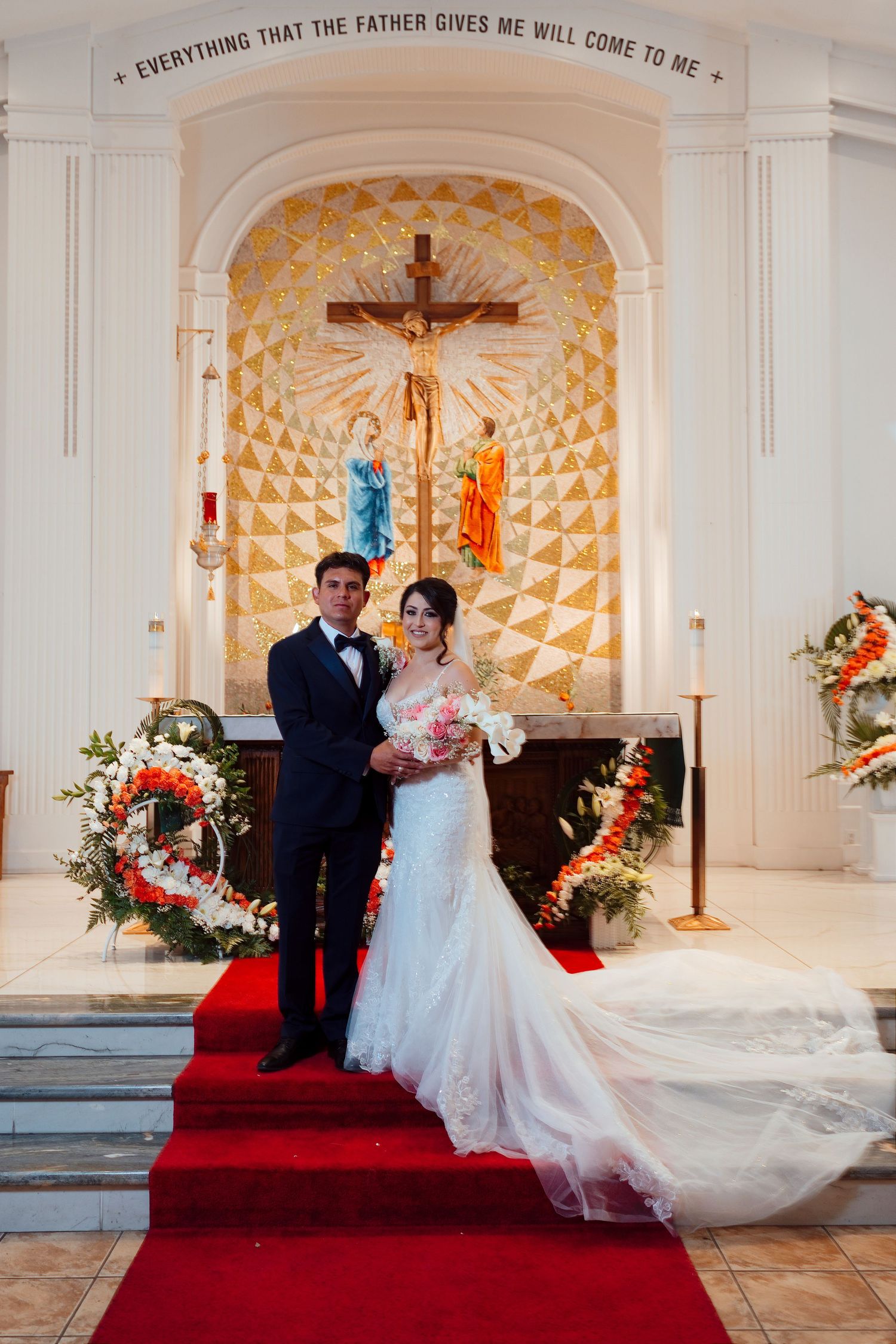 Bride and groom pose at church altar with crucifix and floral decorations on red carpet aisle.