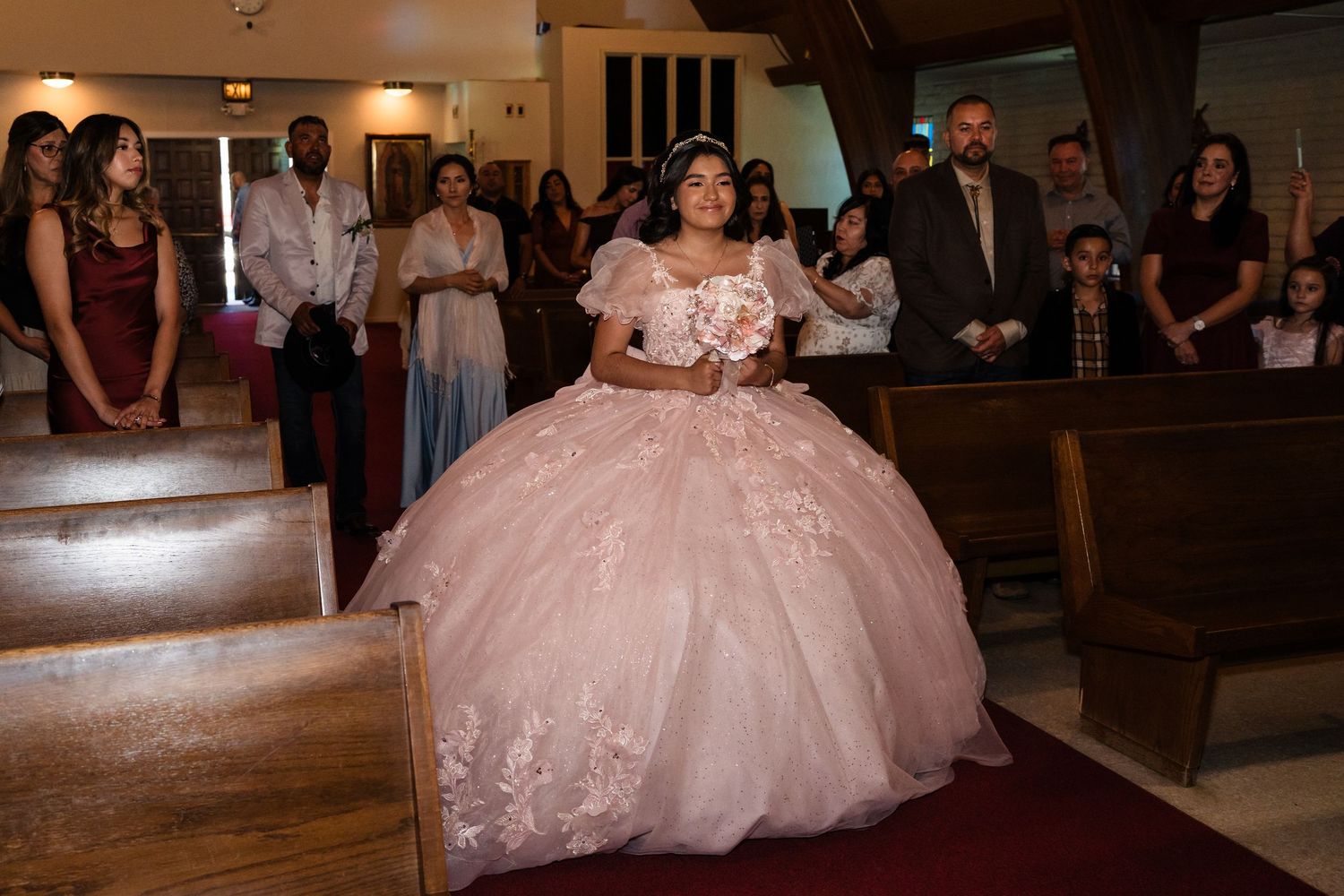 Young girl in elaborate pink ball gown walks down church aisle at her quinceañera.