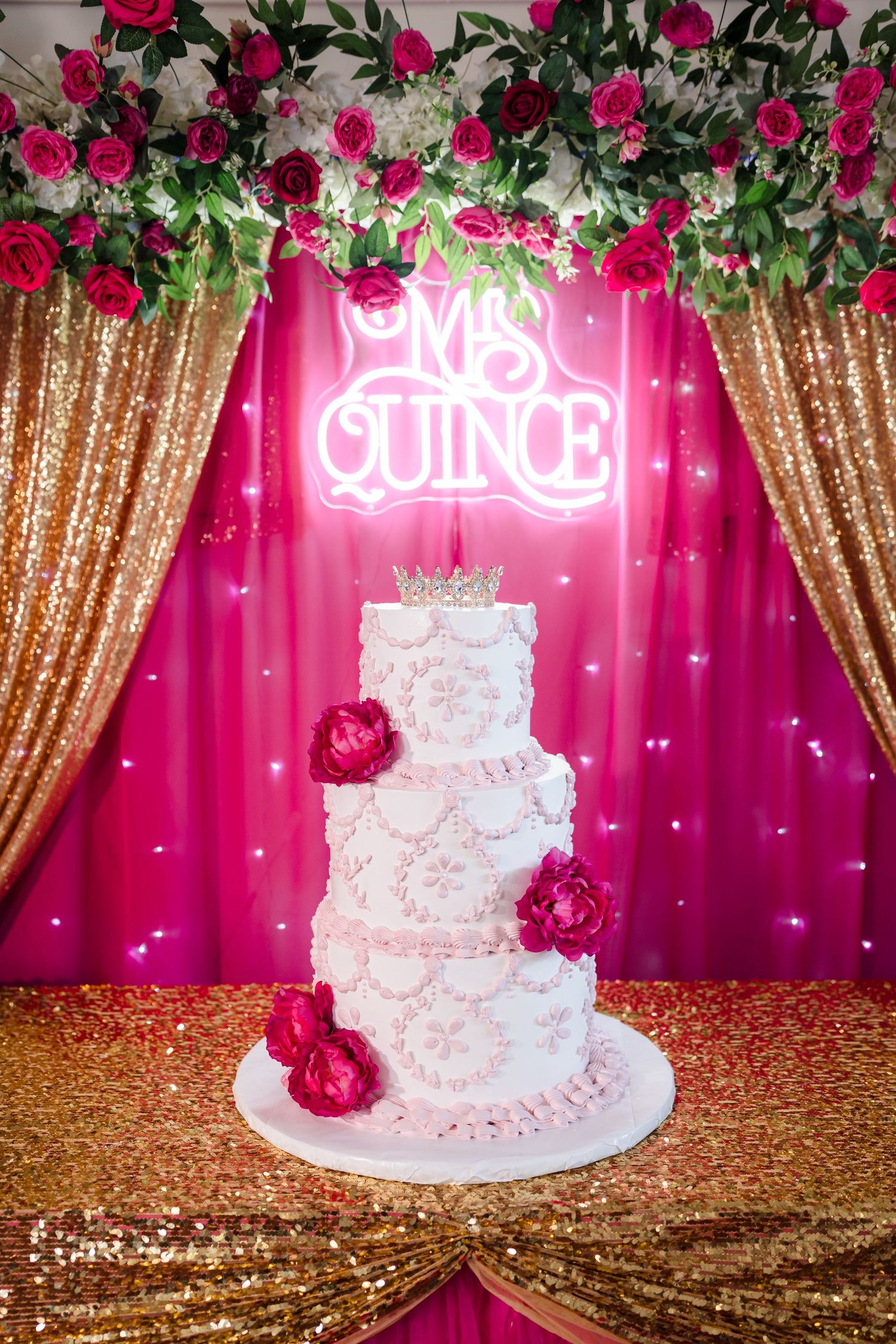 Elegant three-tier white Quinceañera cake with pink peonies, gold sequin tablecloth, and glowing neon sign backdrop.