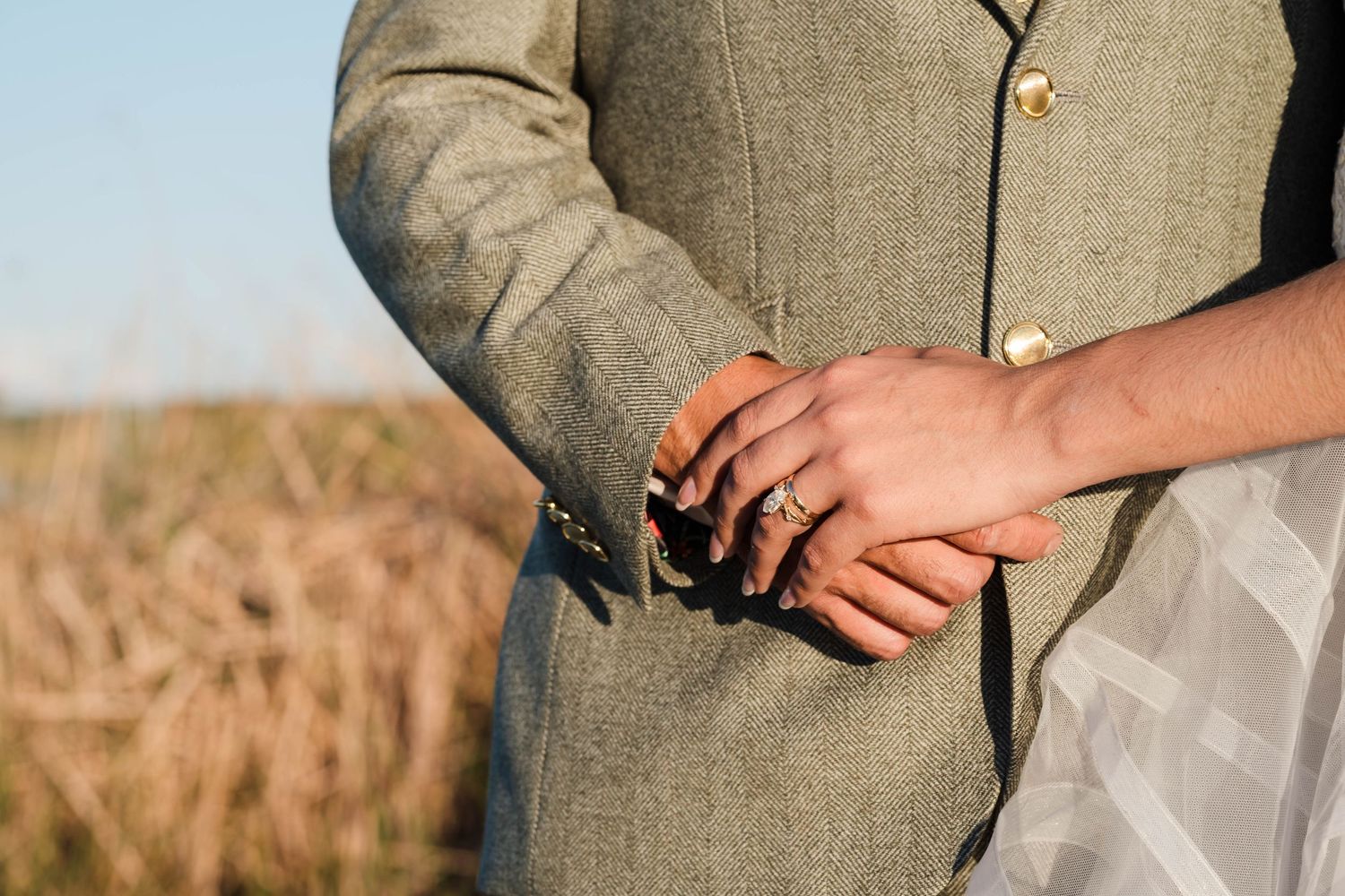 Close-up of a couple holding hands outdoors, wearing wedding rings, with a field in the background.