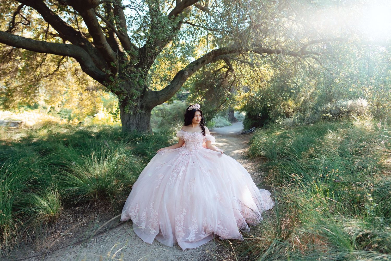 Young woman in a pink ball gown quinceañera dress posing on a garden path beneath a large oak tree.