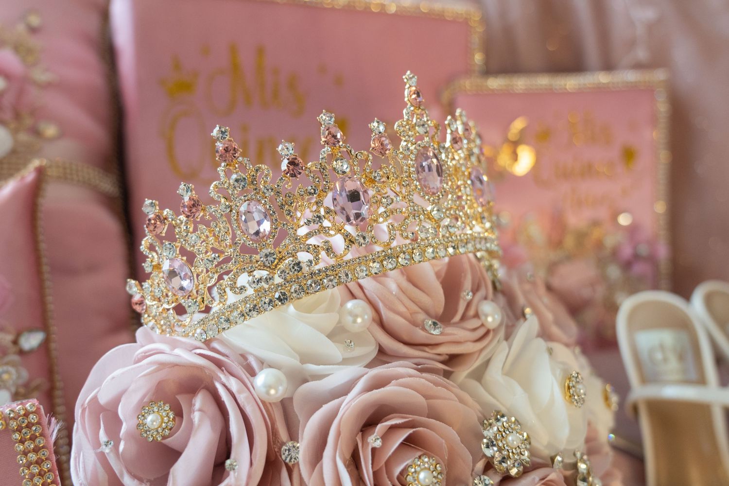 Gold crystal tiara resting on pink fabric roses with pearl accents, elegant quinceañera party decorations in blush pink.
