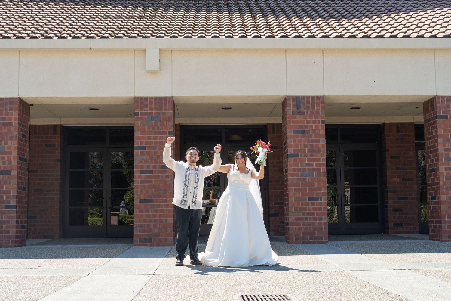 Joyful newlyweds celebrate outside a brick building, raising their hands in triumph in wedding attire.