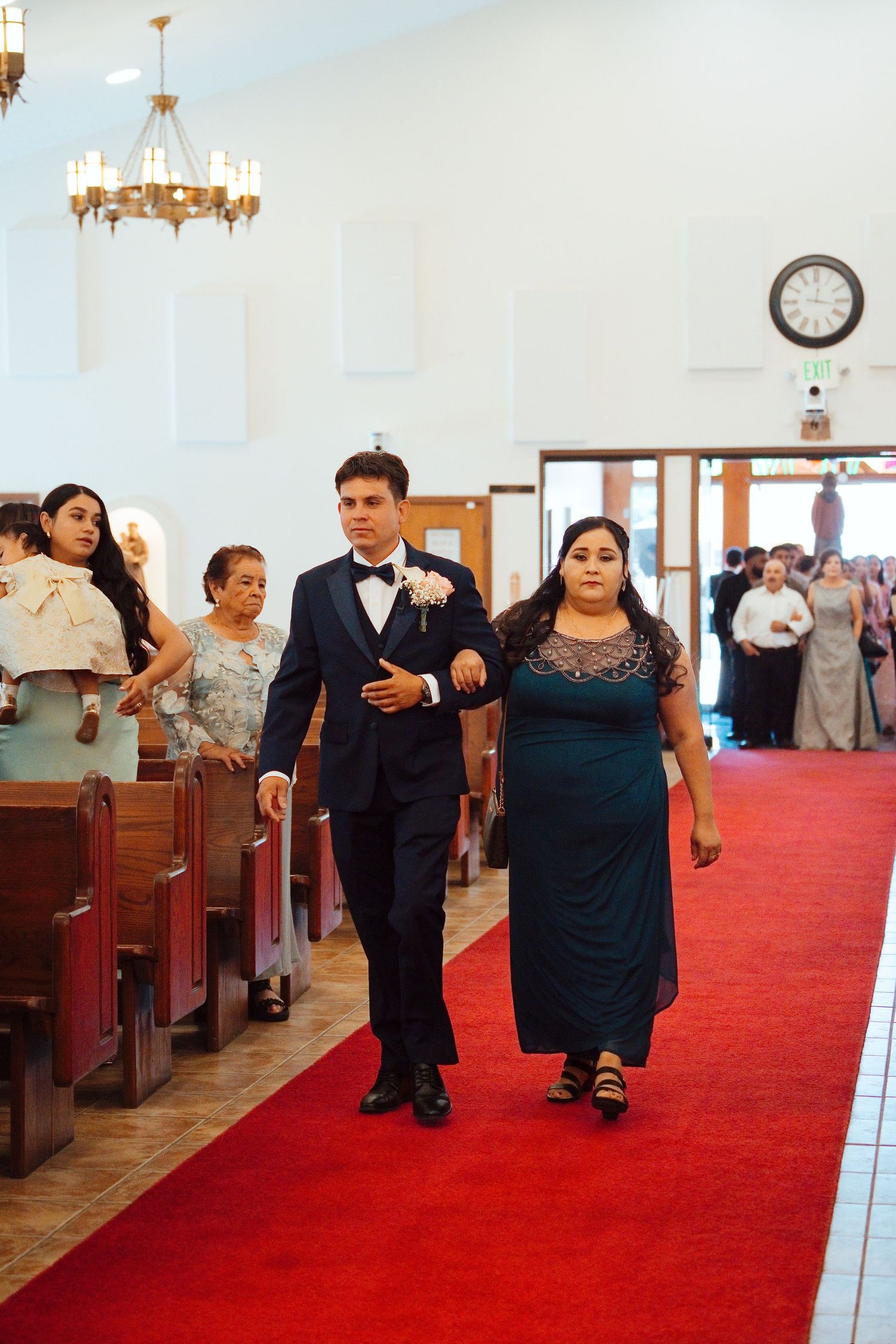 Groom in navy suit escorts woman in teal dress down red carpet aisle in church wedding ceremony.