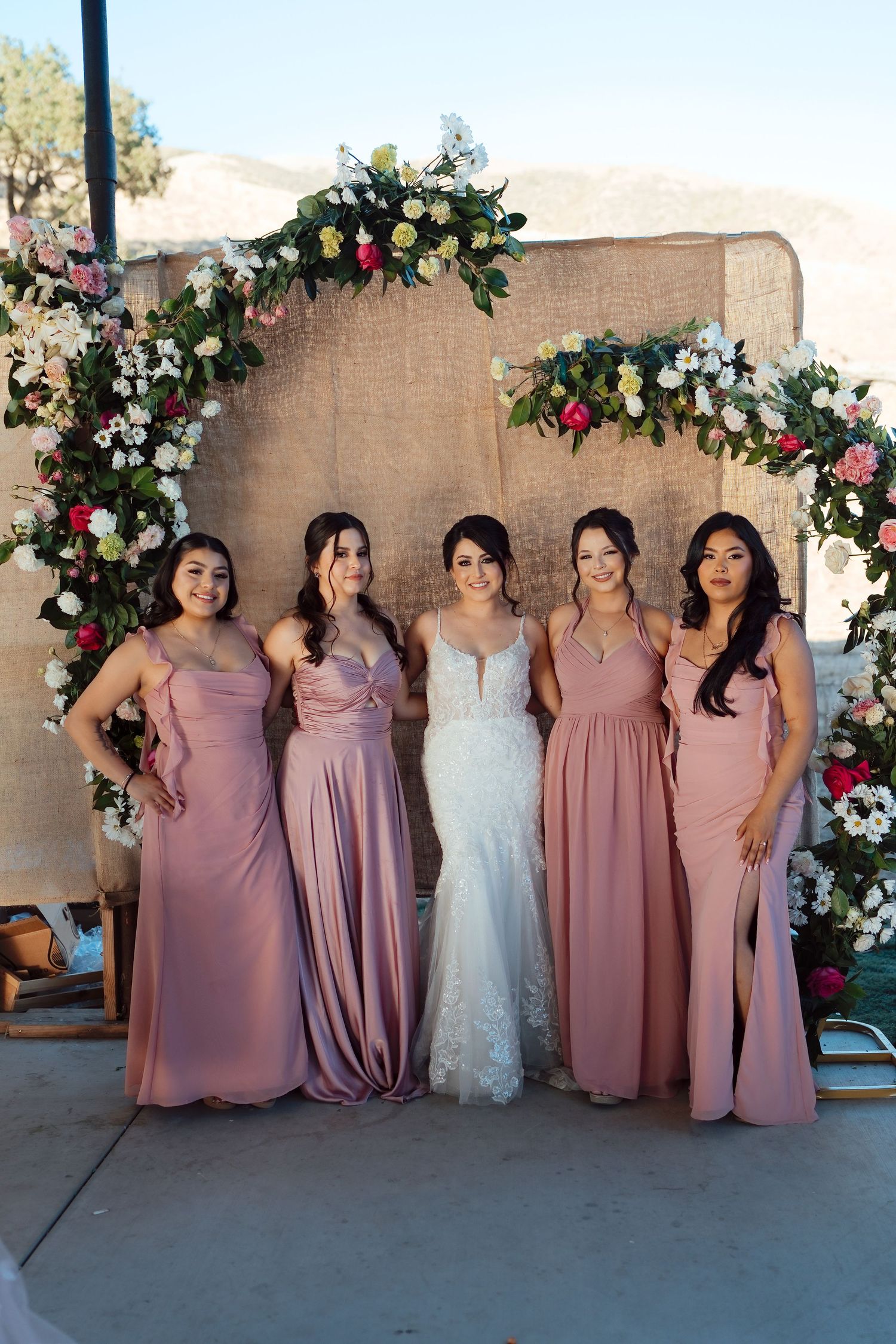Bride in white lace gown poses with four bridesmaids in pink dresses before a floral burlap arch.