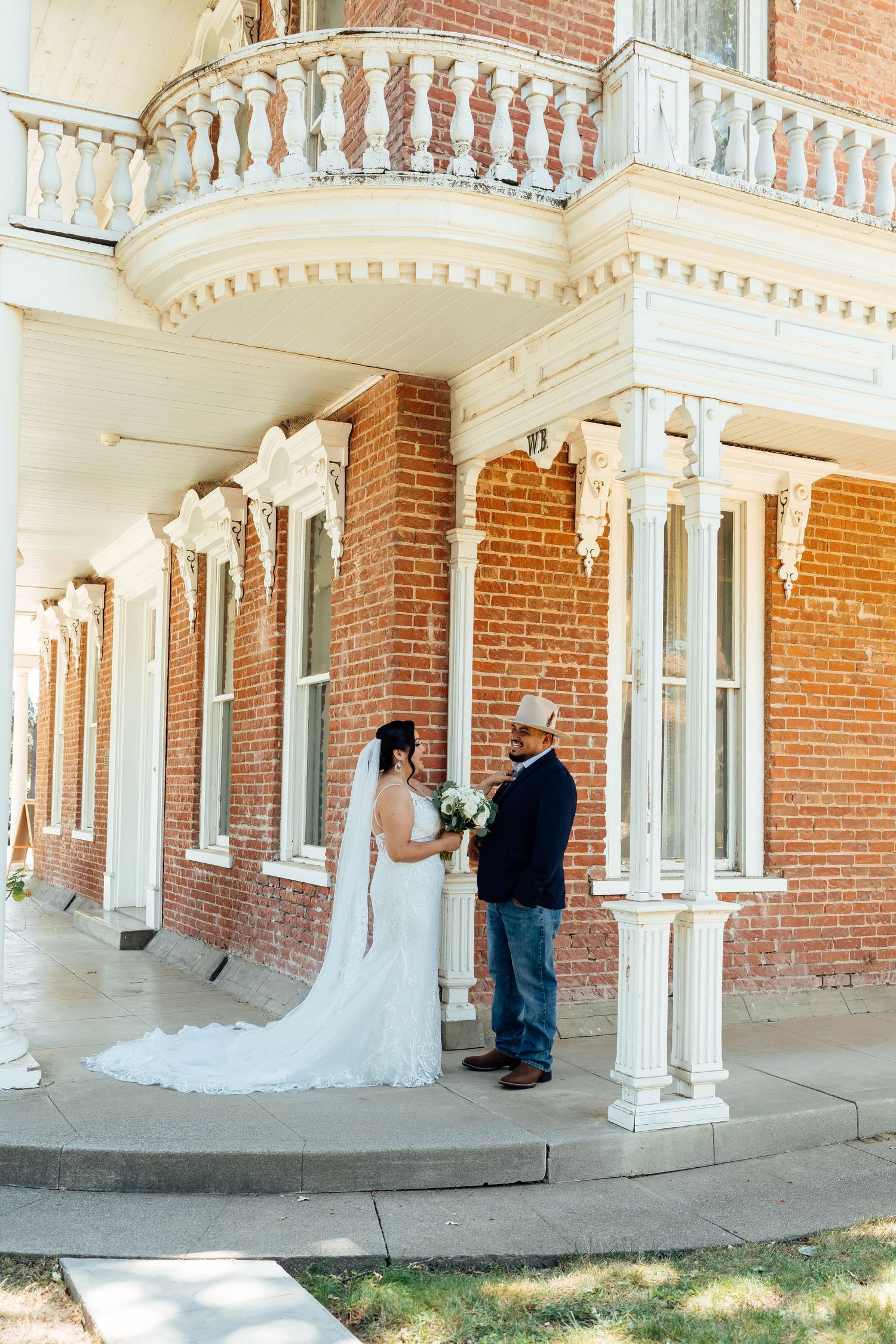 Wedding couple stands together in front of a historic brick mansion with ornate white balcony.