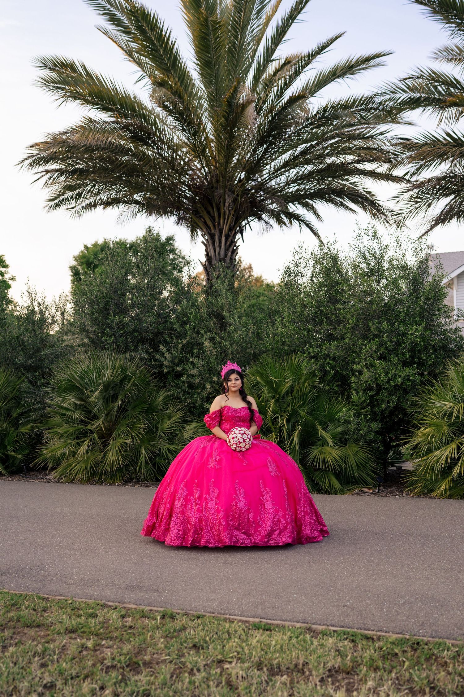 Teen in hot pink quinceañera ball gown with bouquet poses before large tropical palm tree.