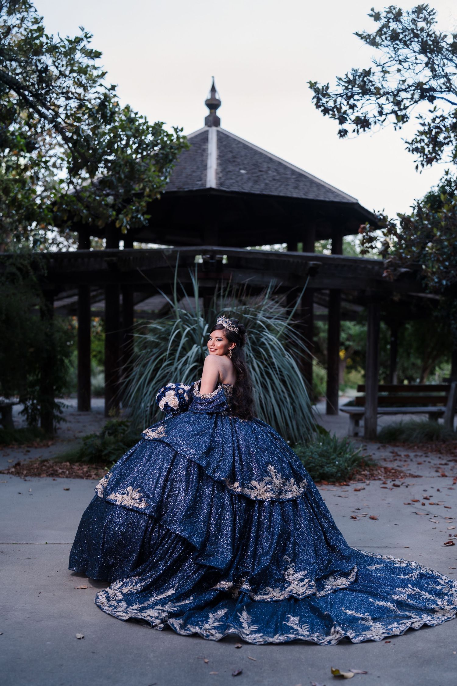 Young woman in a navy blue sequined quinceañera gown with gold embroidery posing near a park gazebo.