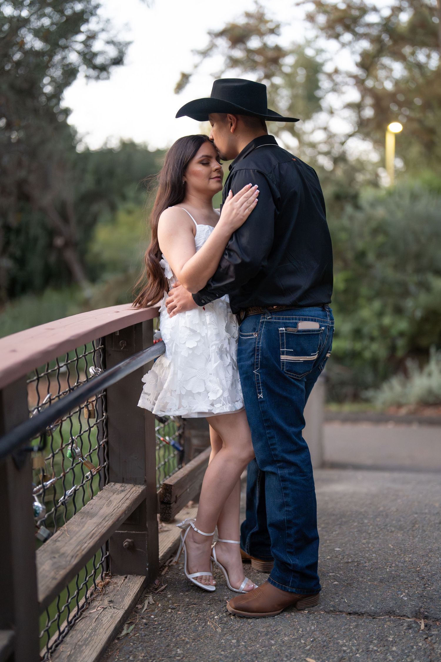 Couple sharing a kiss on a bridge, man in black cowboy hat, woman in white dress with silver heels.