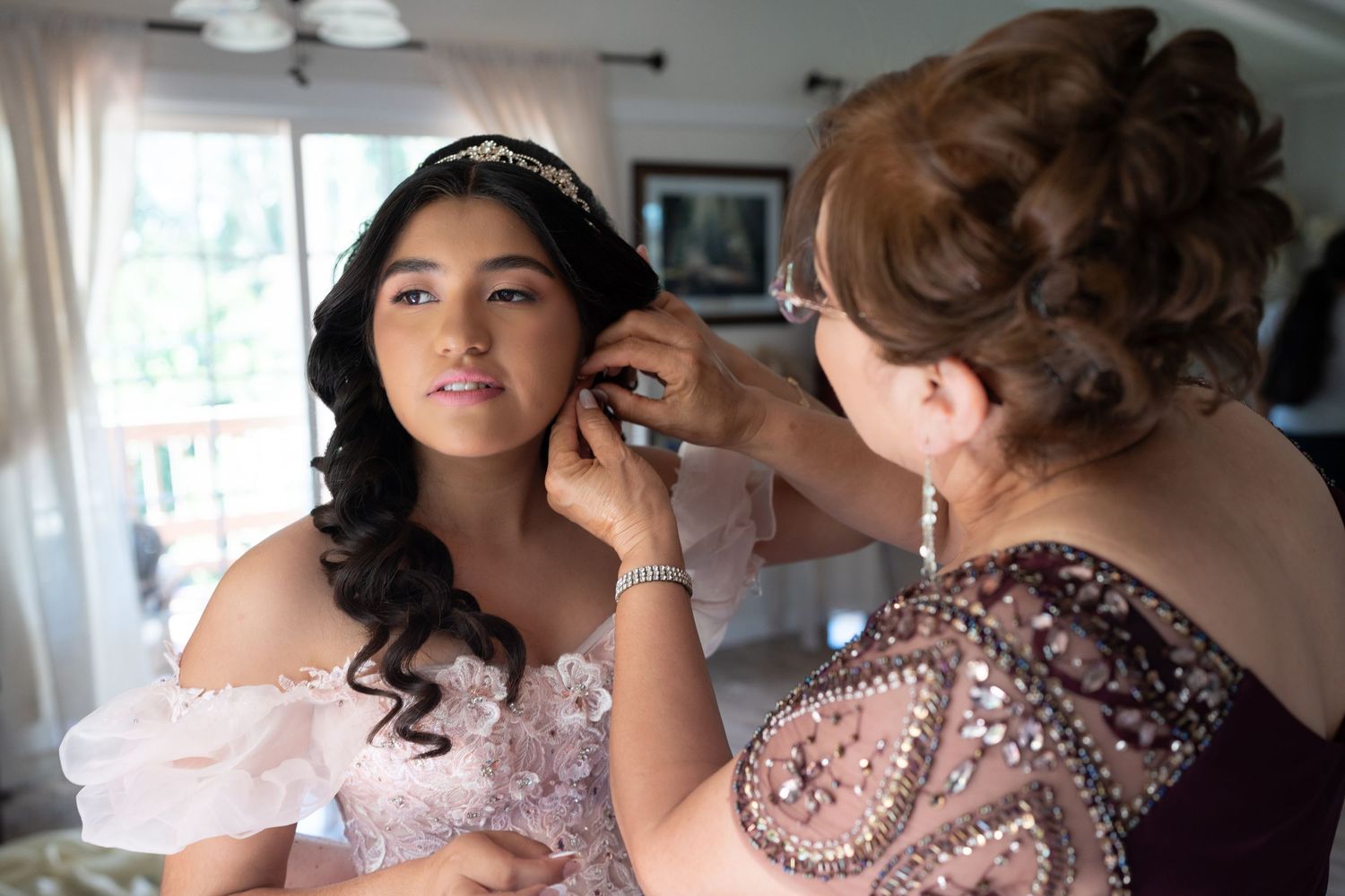 A woman helps a young Quinceañera adjust her earrings while wearing a pink gown and tiara.