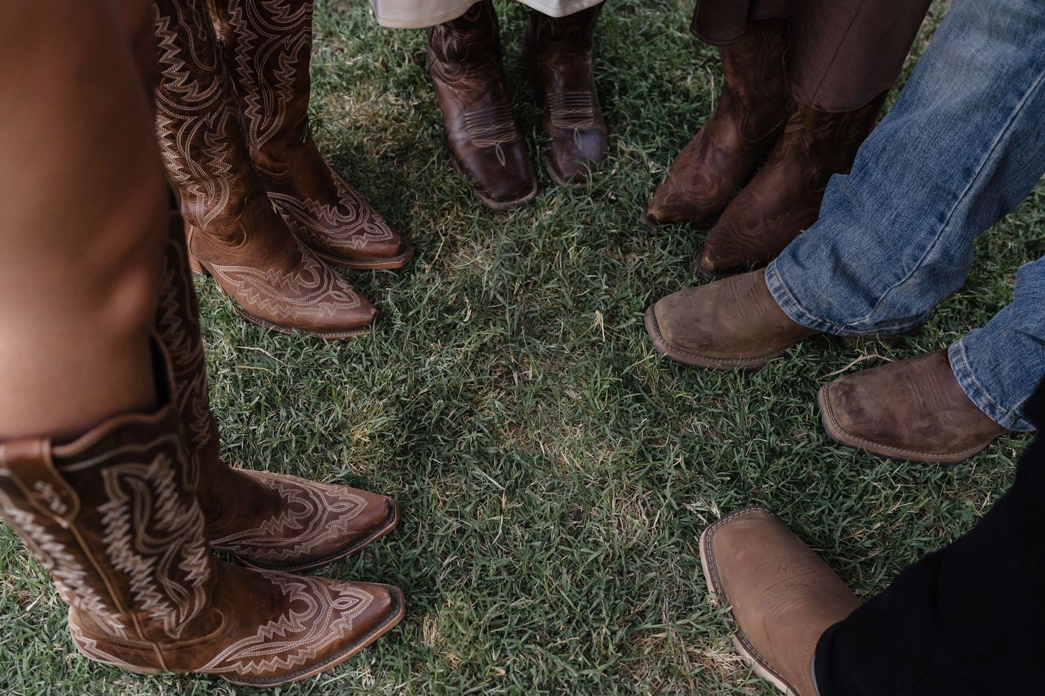 Group of people wearing brown cowboy boots standing in a circle on green grass.