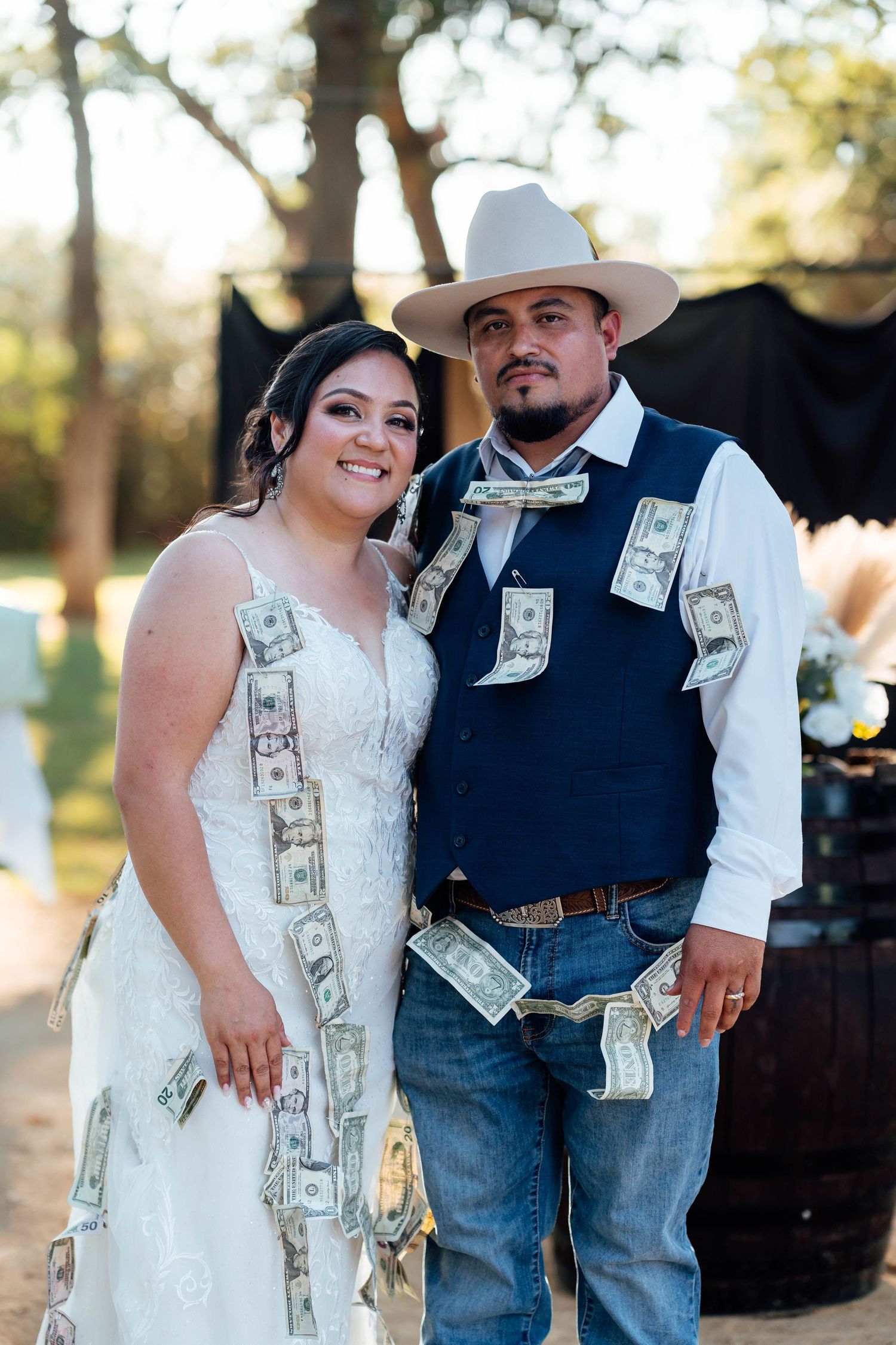 Bride and groom covered in dollar bills pose together smiling at outdoor western-themed wedding reception.