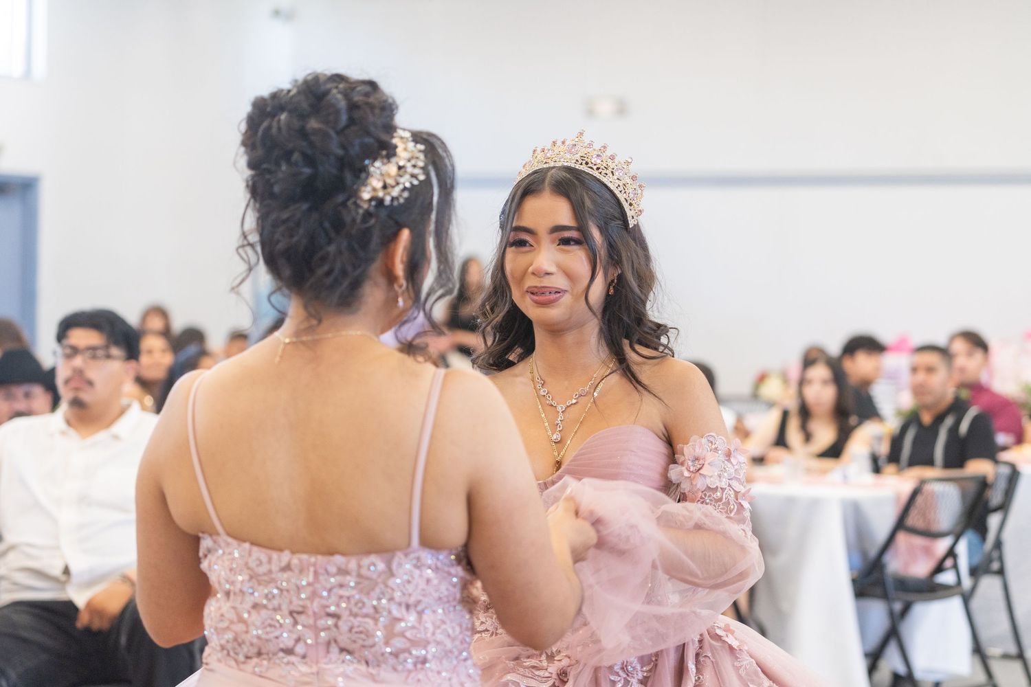 Two young women in pink quinceañera dresses sharing a moment during a celebration reception.