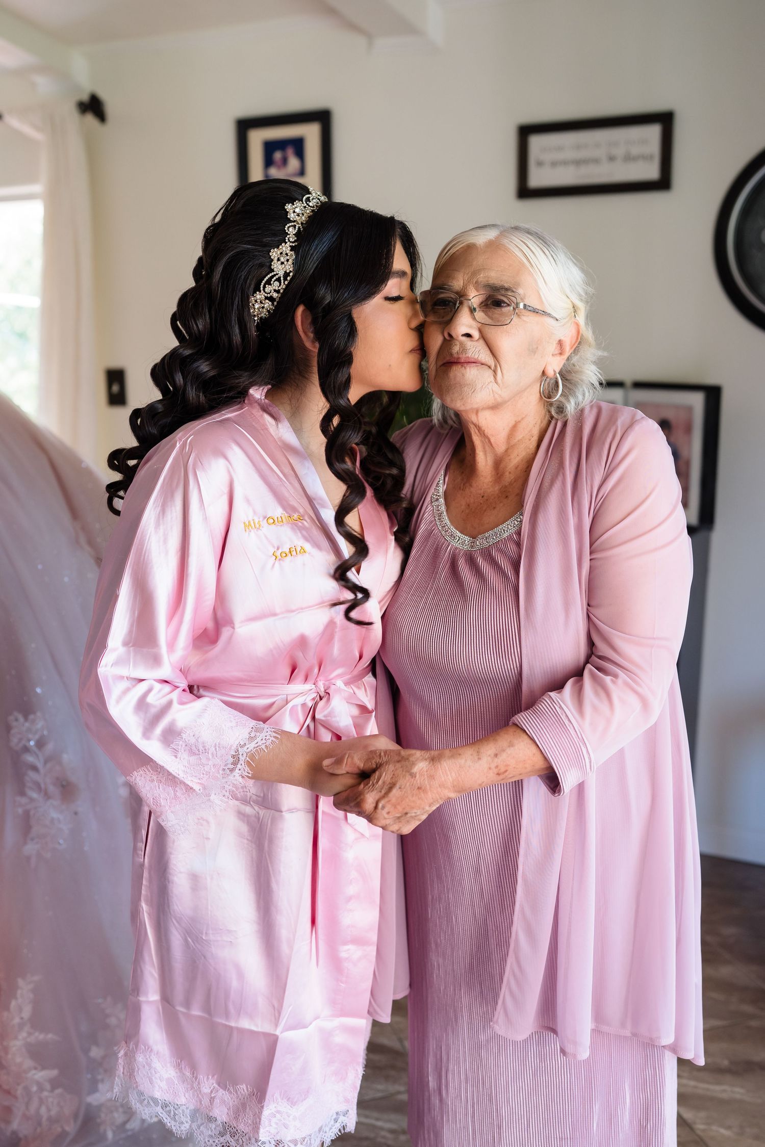 Quinceañera girl in pink robe kisses elderly grandmother in matching pink dress.