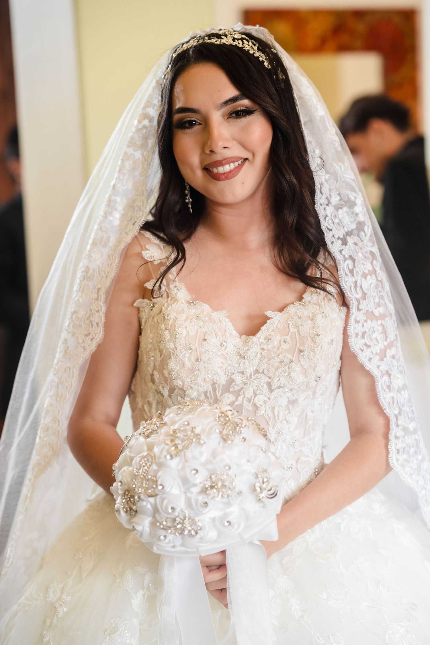 Beautiful bride smiles wearing lace ball gown wedding dress with gold tiara veil and floral bouquet.