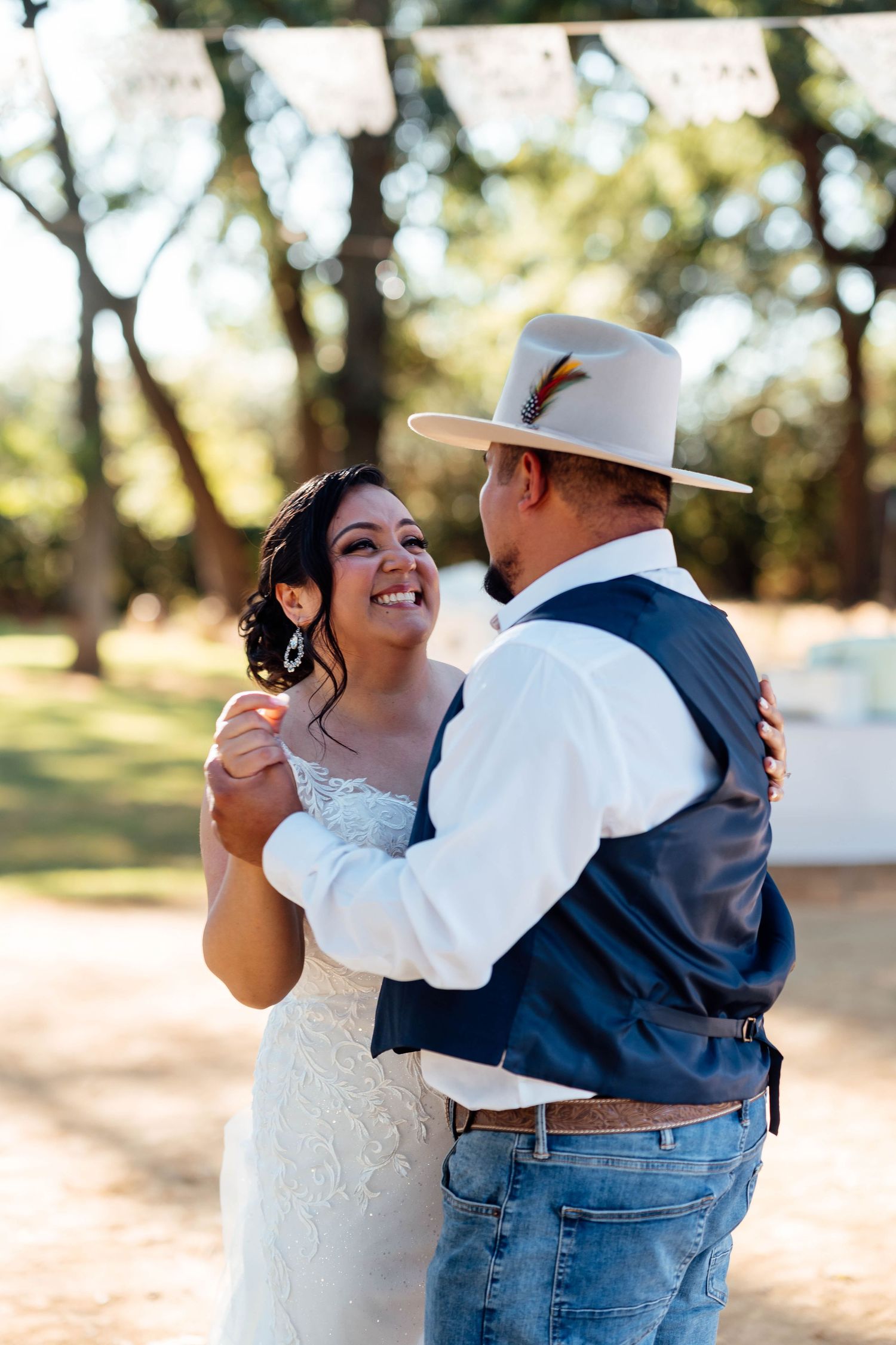 Bride in lace gown smiles joyfully while sharing first dance with groom in cowboy hat and navy vest outdoors.