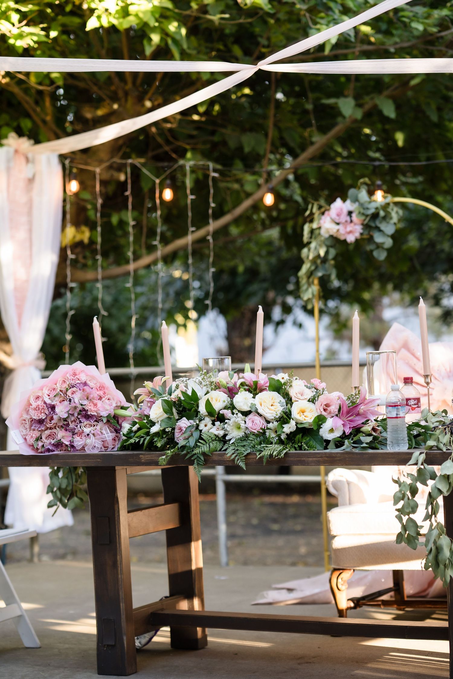 Elegant outdoor wedding reception table adorned with pink and white floral arrangements, candles, and greenery under string lights.