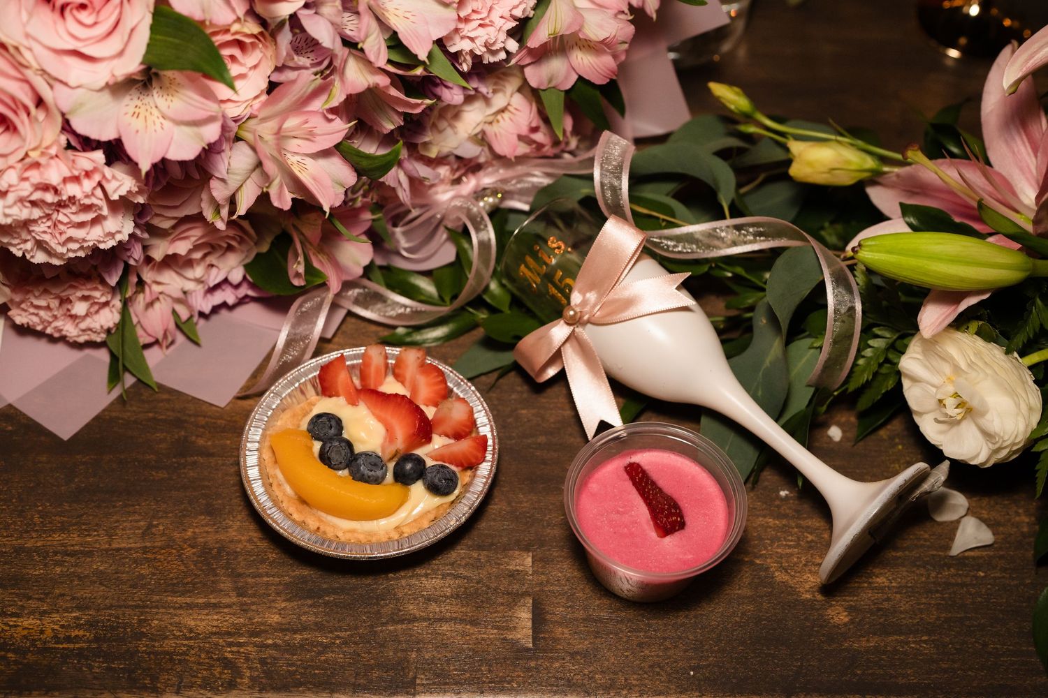 Elegant celebration setup with fruit tart, pink champagne, and fresh floral bouquet on wooden table.