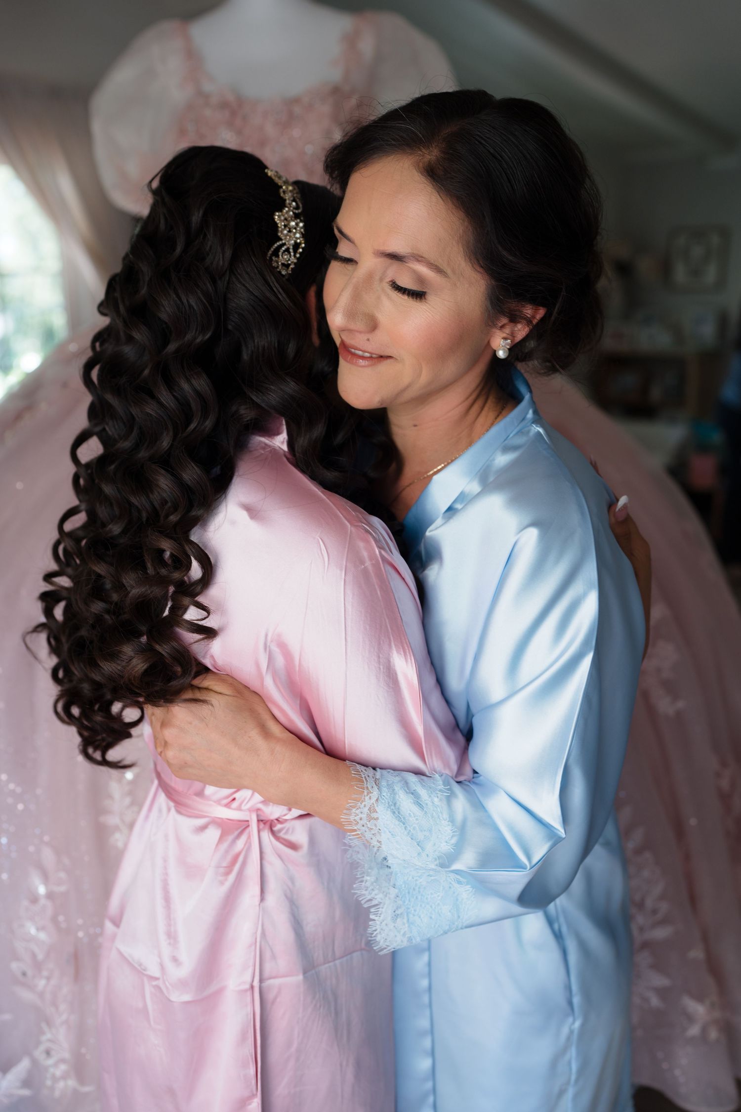 Two women in satin robes share a warm embrace during quinceañera preparations.