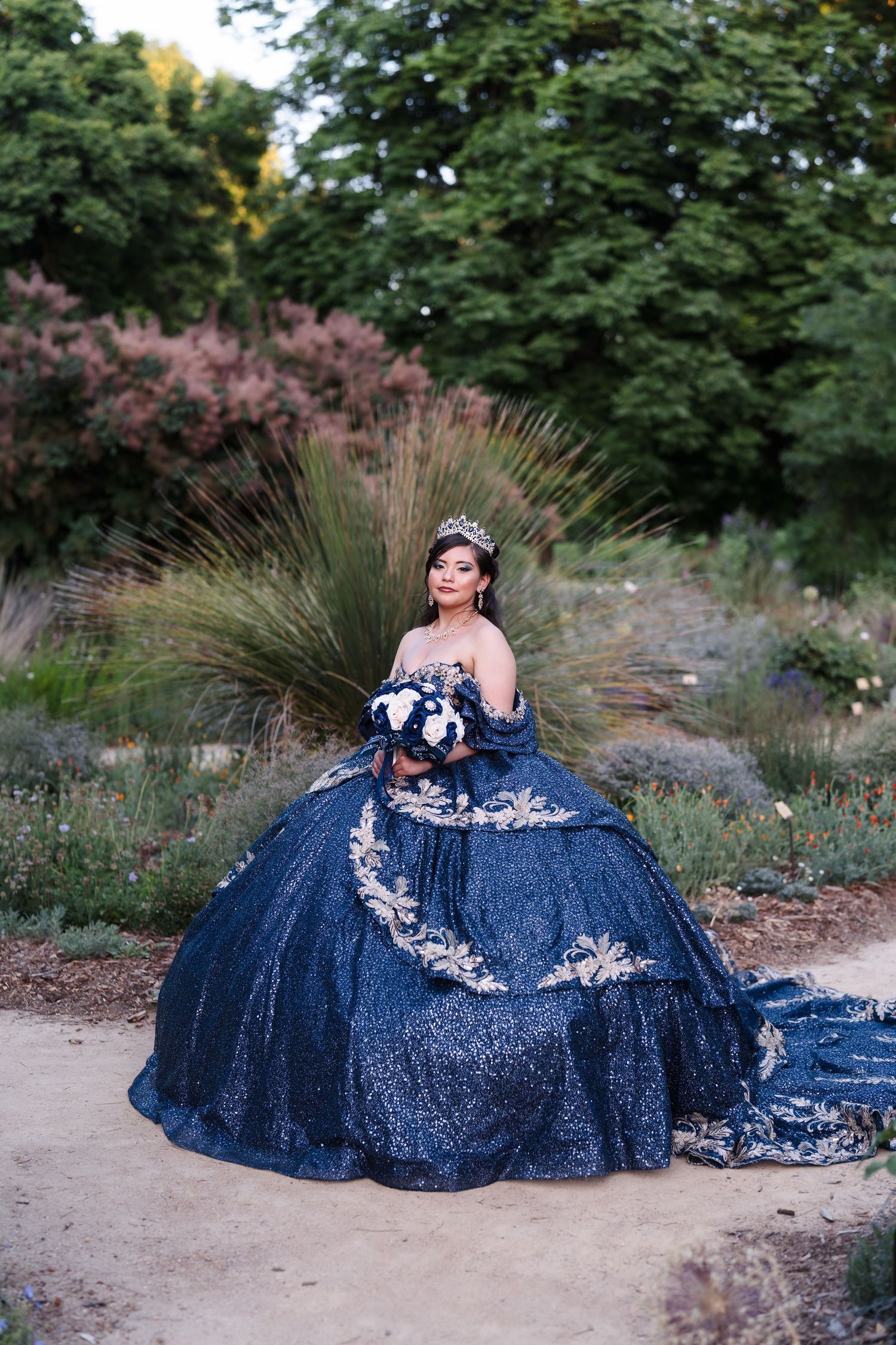 Young woman in navy blue sequined quinceañera ballgown with gold lace, tiara, and floral bouquet in garden setting.