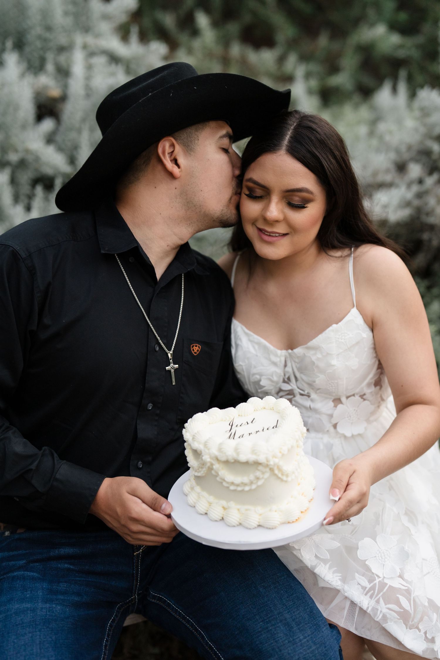 Groom in black cowboy hat kissing bride on cheek while she holds a Just Married white wedding cake.