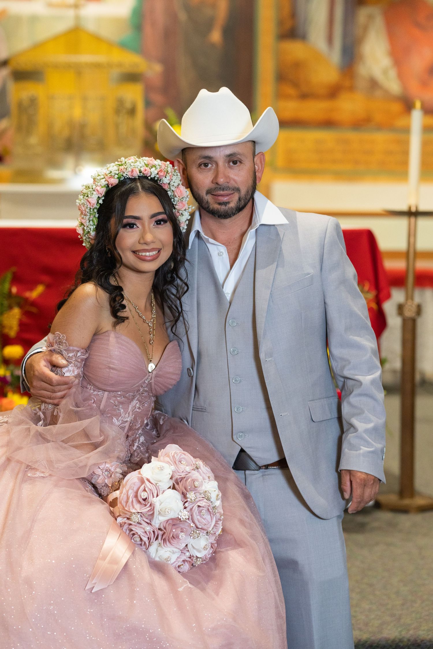 Quinceañera girl in pink ball gown with floral crown posing with man in gray suit and white cowboy hat.