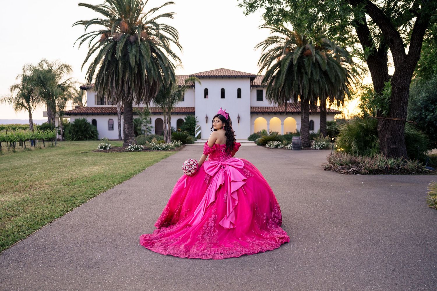 A young woman in a vibrant hot pink quinceañera gown poses on a pathway before a grand estate.