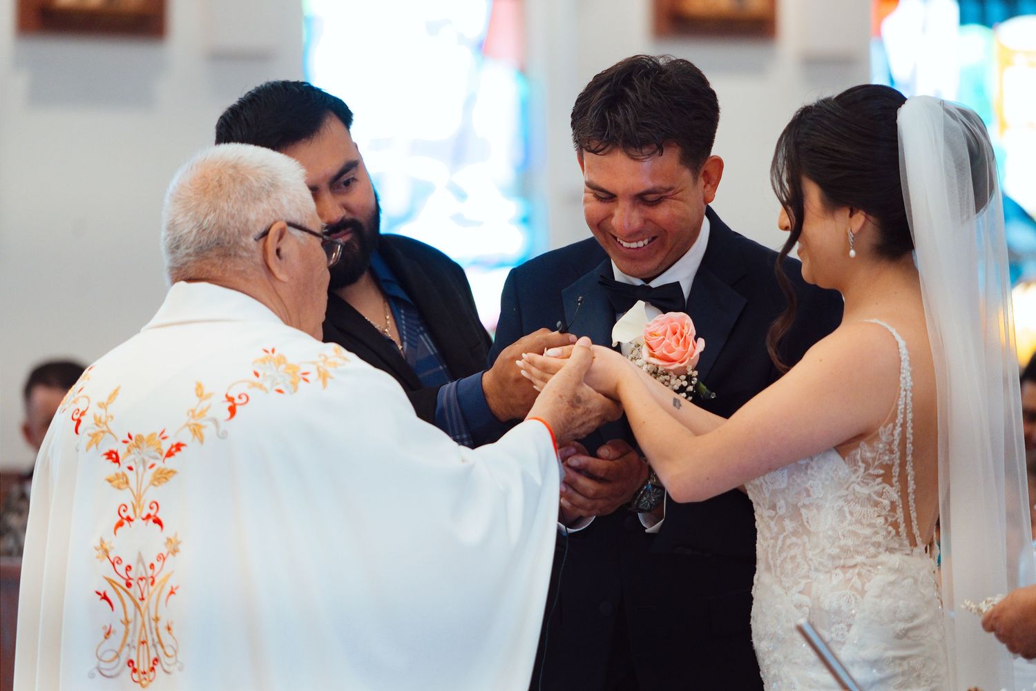 Priest officiating wedding ring exchange ceremony with smiling groom and bride in white dress inside a church.
