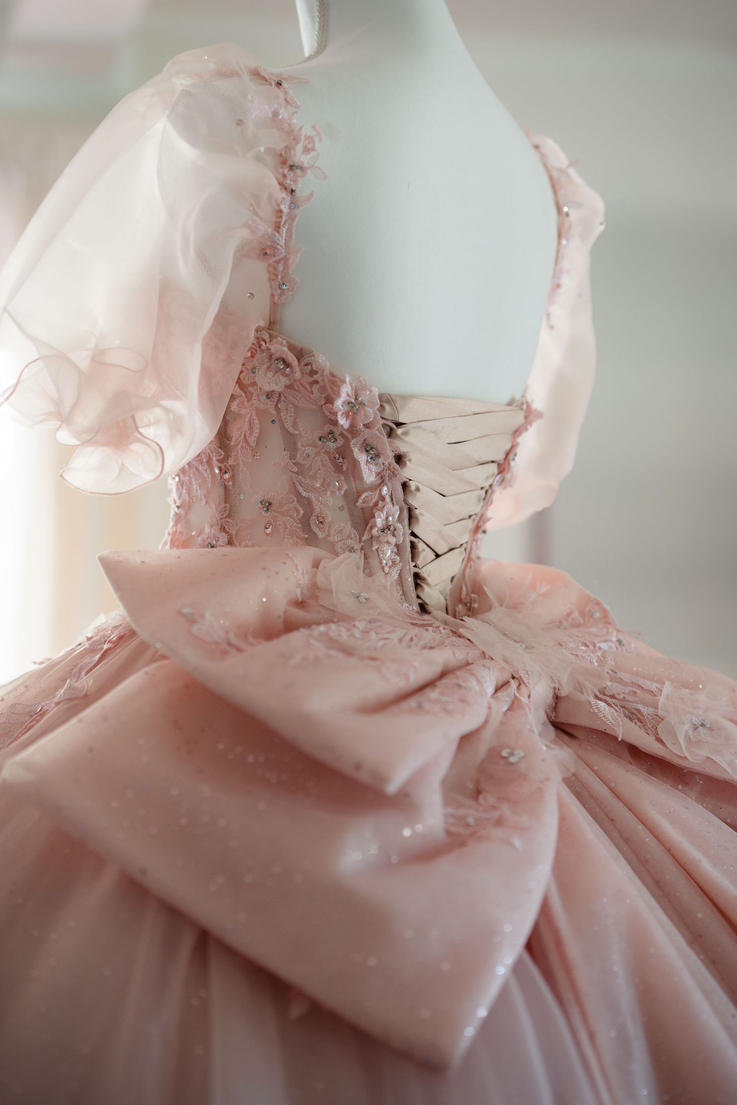 Elegant blush pink ball gown with corset back, lace details, puff sleeves, and large bow on a mannequin.