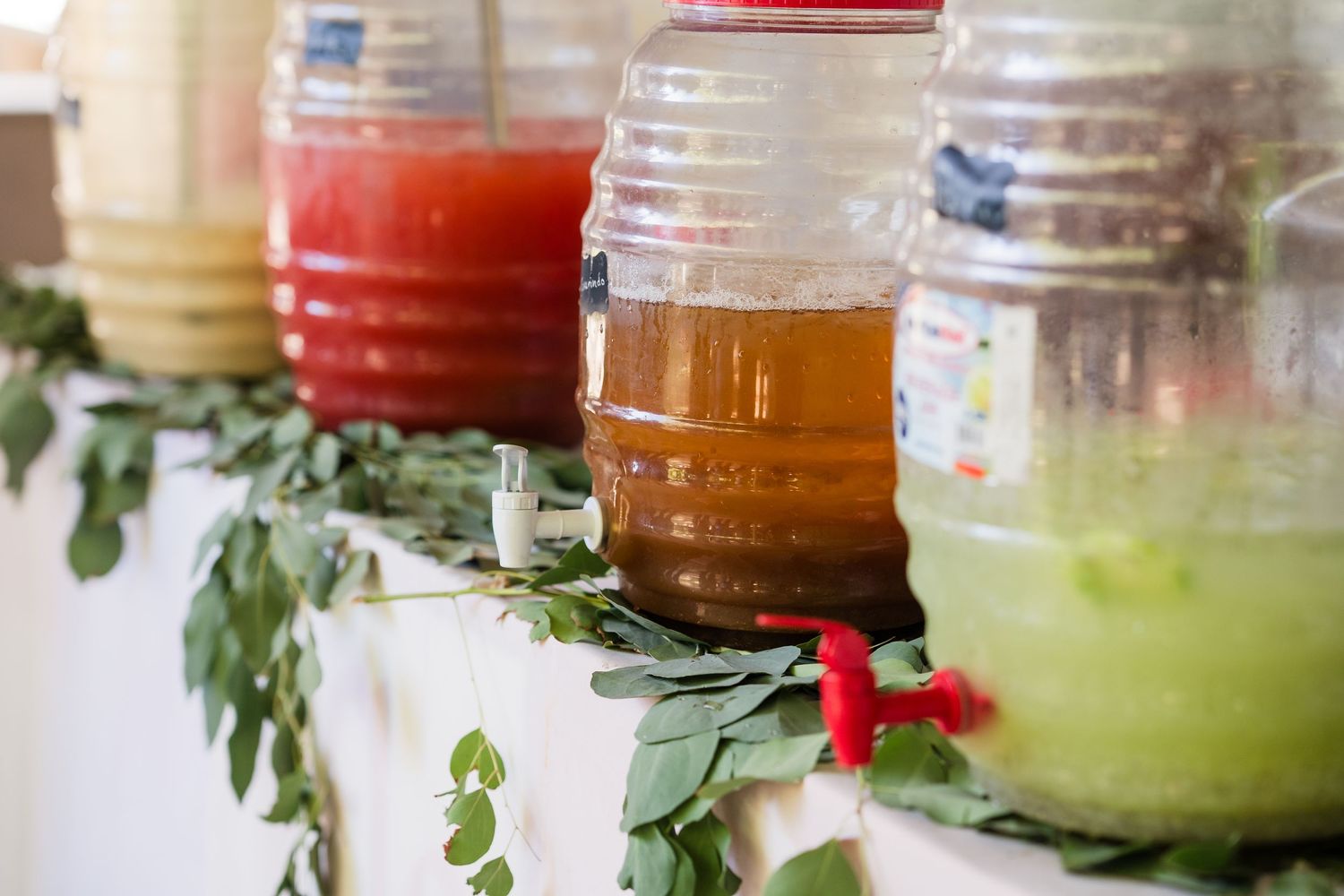 Colorful beverage dispensers with spigots lined up on a white shelf decorated with fresh green garland.