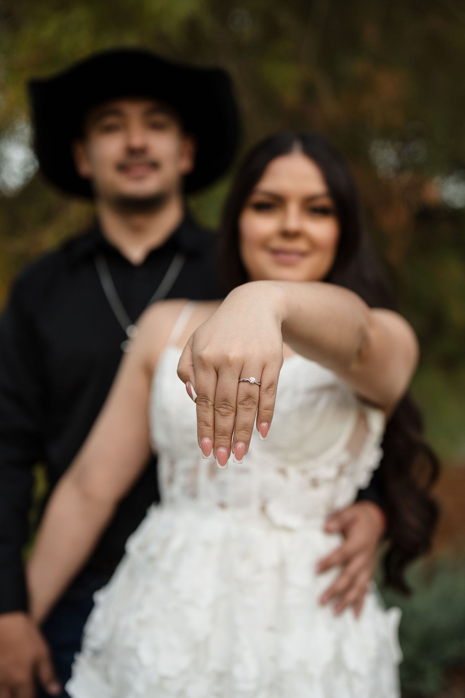 Bride extending hand toward camera showing engagement ring, groom in black cowboy hat standing behind her.