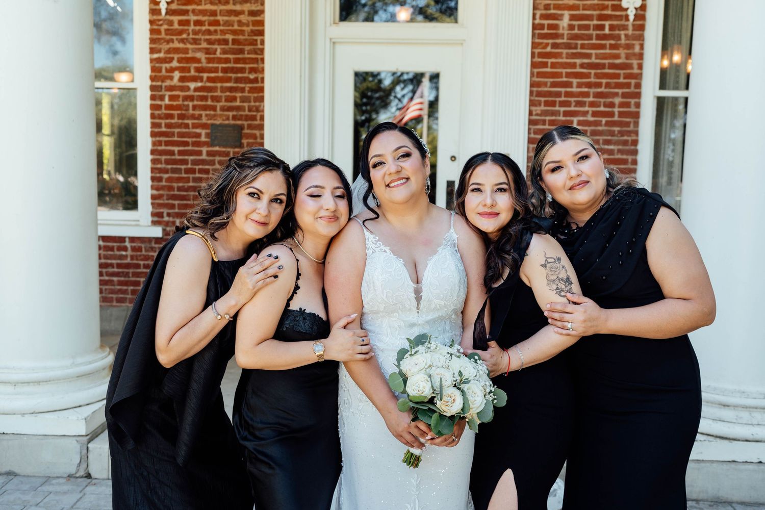 Bride surrounded by four bridesmaids in black dresses, all smiling in front of a brick building.