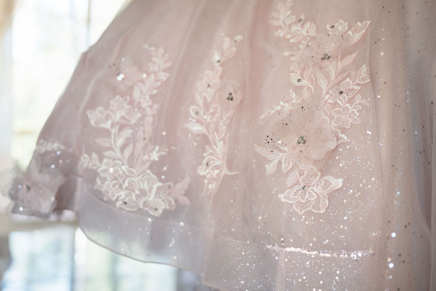 Close-up of a blush pink wedding dress hem with white floral lace appliques and sparkling sequin embellishments.