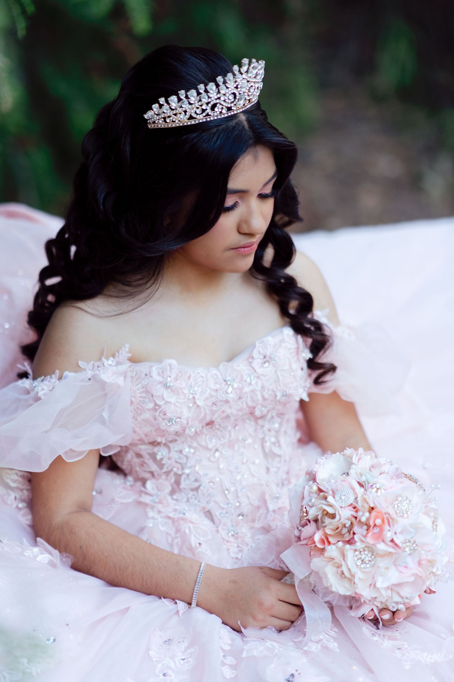 Young woman in pink off-shoulder quinceañera gown with crystal tiara holds floral bouquet outdoors.