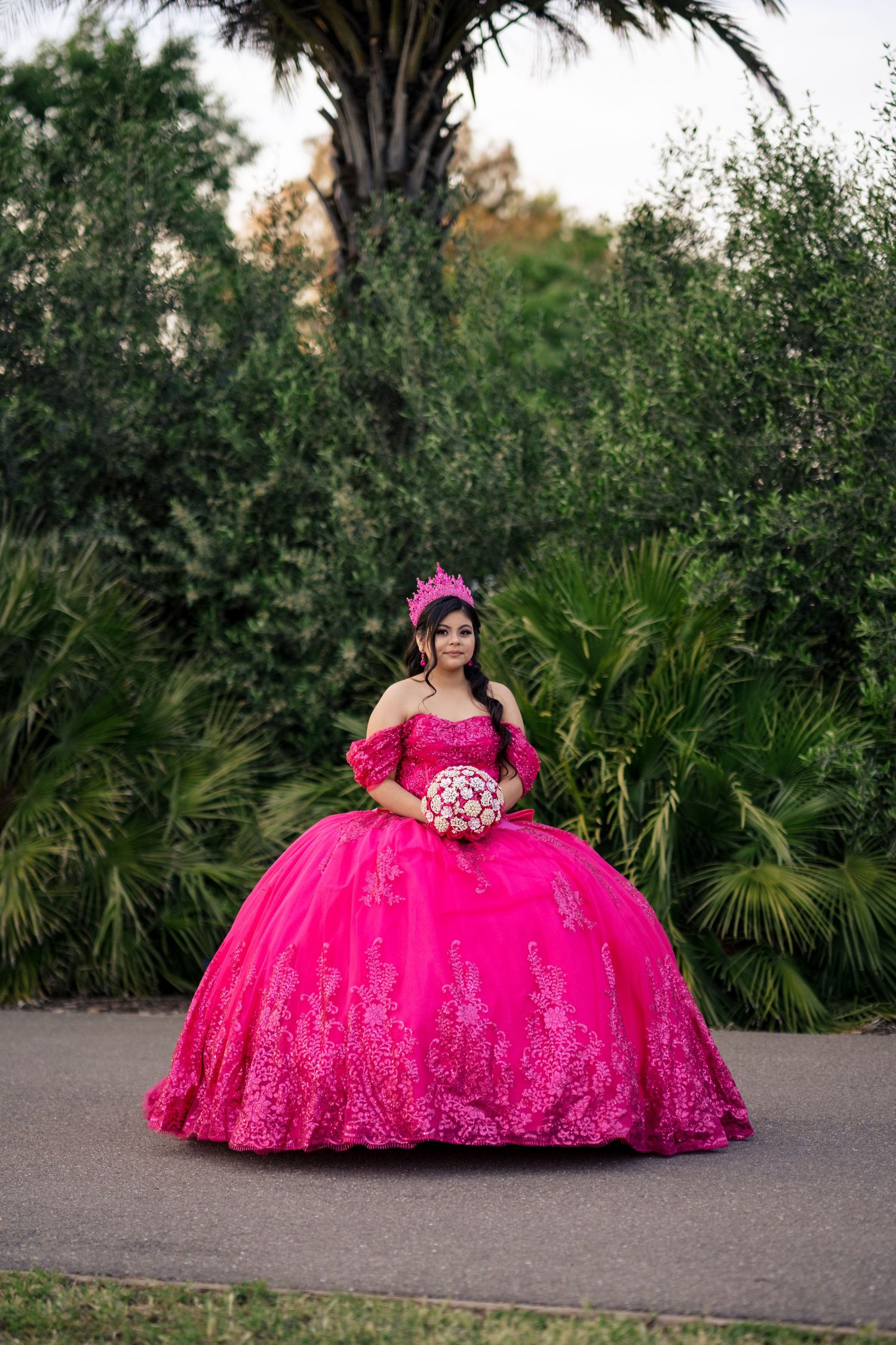 Young woman in full hot pink quinceañera gown with crown holds bouquet on garden pathway.